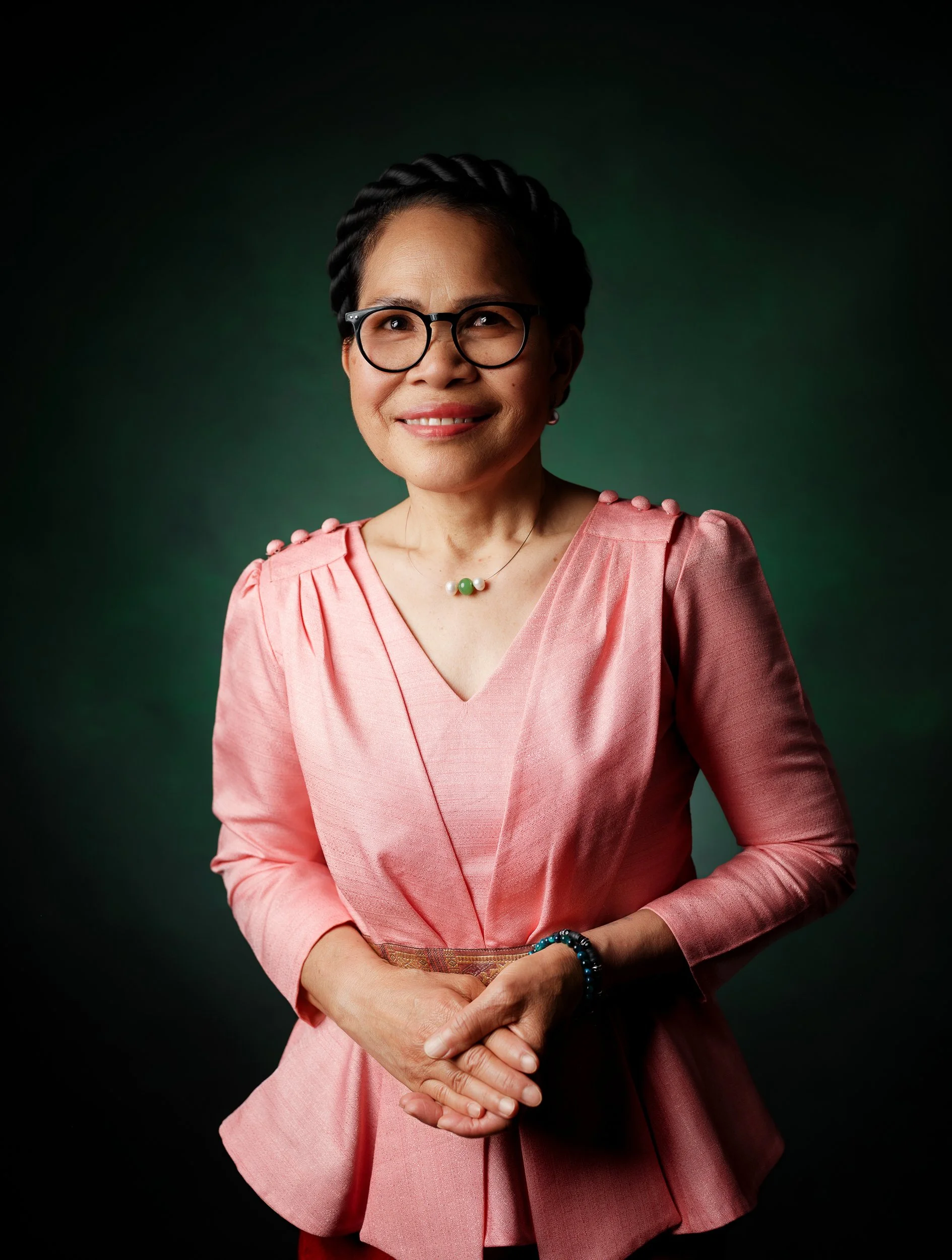 Porträt einer lachenden Frau mit Brille, Kurzhaarfrisur, in einem pinkfarbenen Blazer, steht vor dunklem Hintergrund.