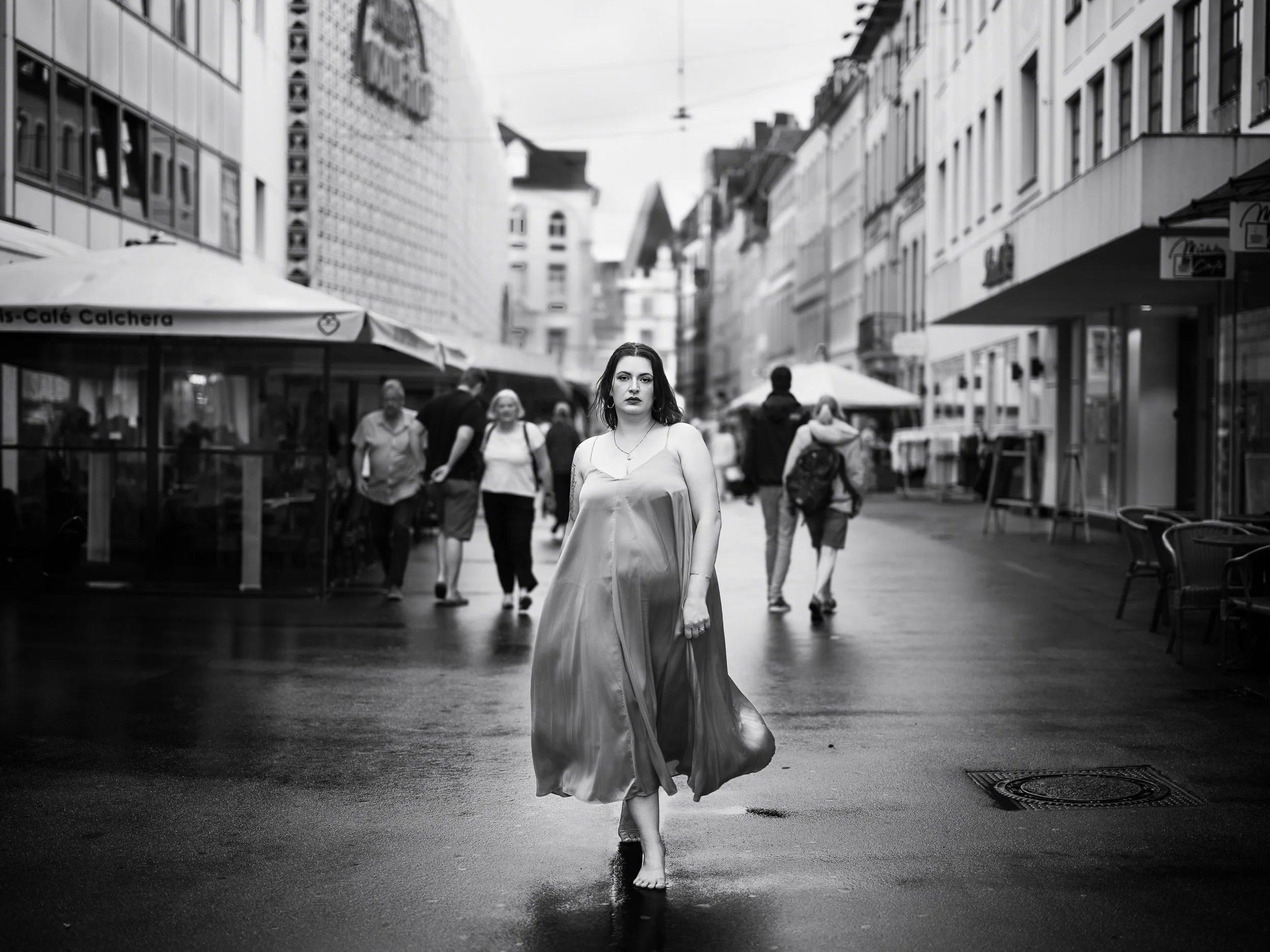 Eine Frau in einem langen Kleid geht barfuß auf einer nassen Stadtstraße, umgeben von vorbeigehenden Menschen, in einer urbanen Umgebung mit Geschäften und Gebäuden.