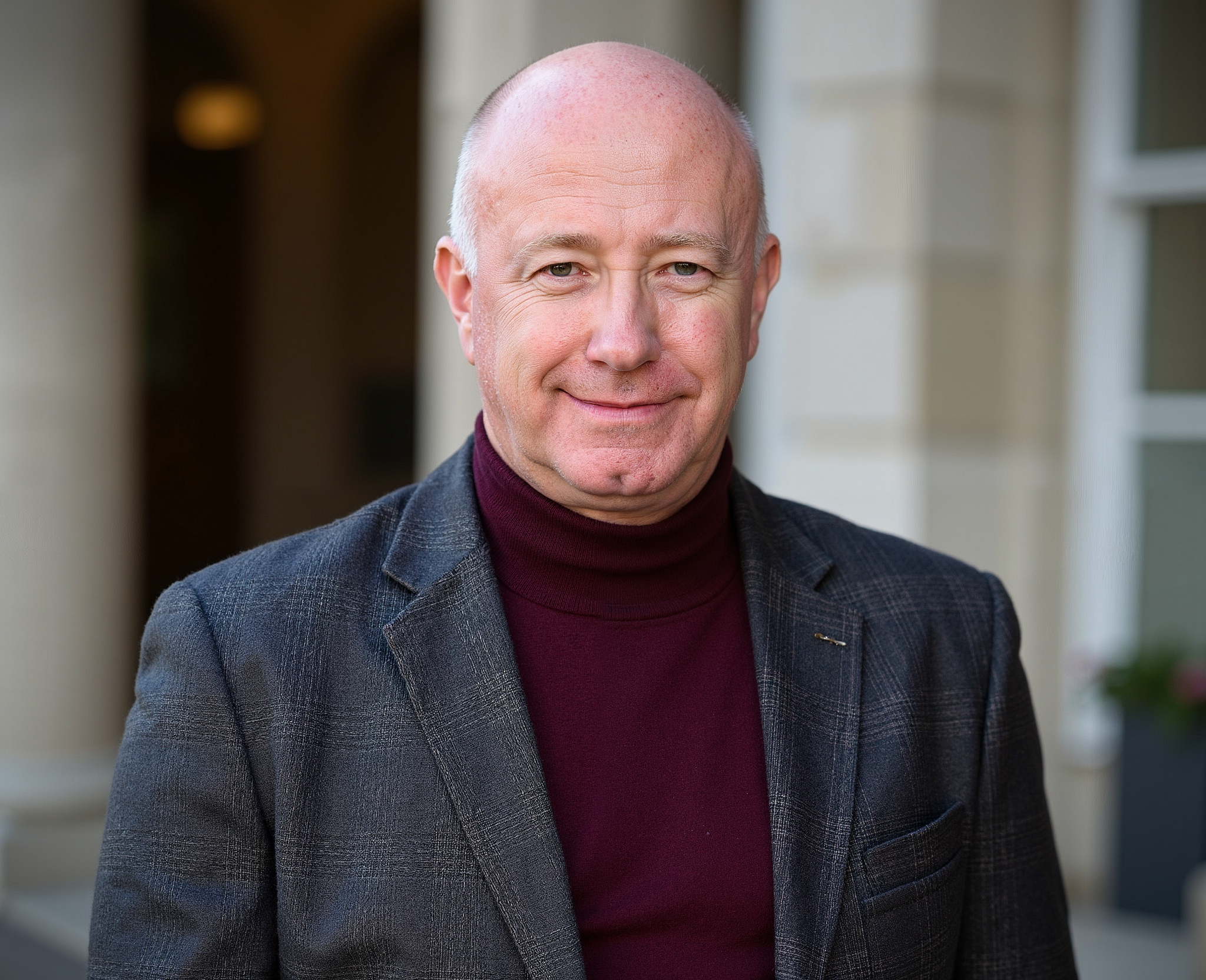 A middle-aged man with a bald head, wearing a dark plaid blazer and a maroon turtleneck, standing outdoors in front of a building with large white columns and windows.