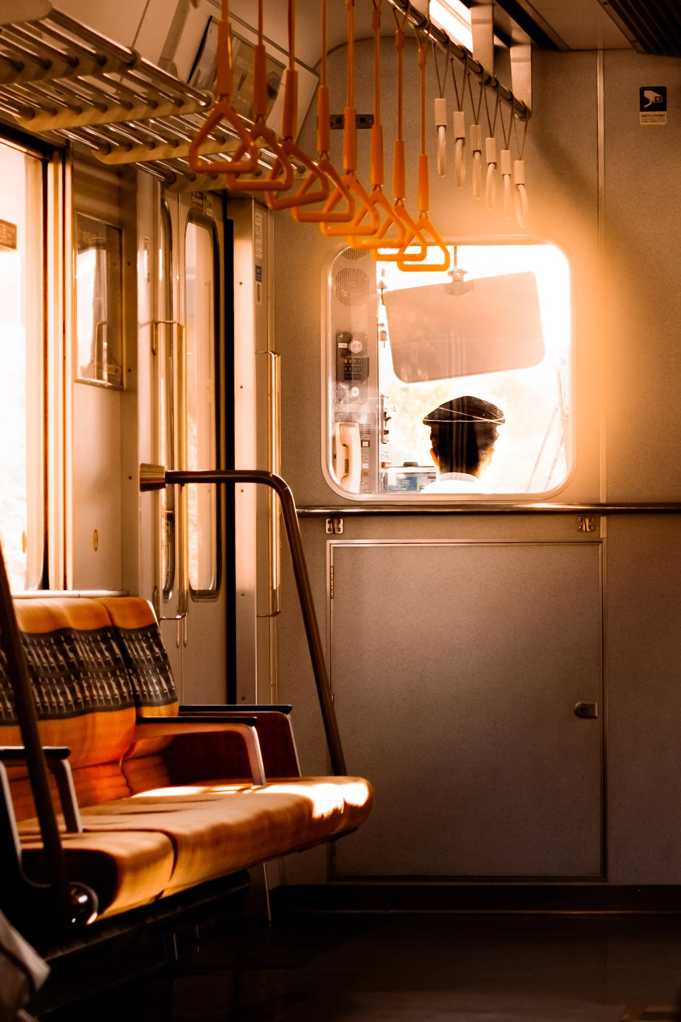
Intérieur d’un train avec sièges orange et jaune vides, poignées de maintien suspendues, lumière du soleil, et silhouette d’un agent ferroviaire visible à travers la fenêtre.