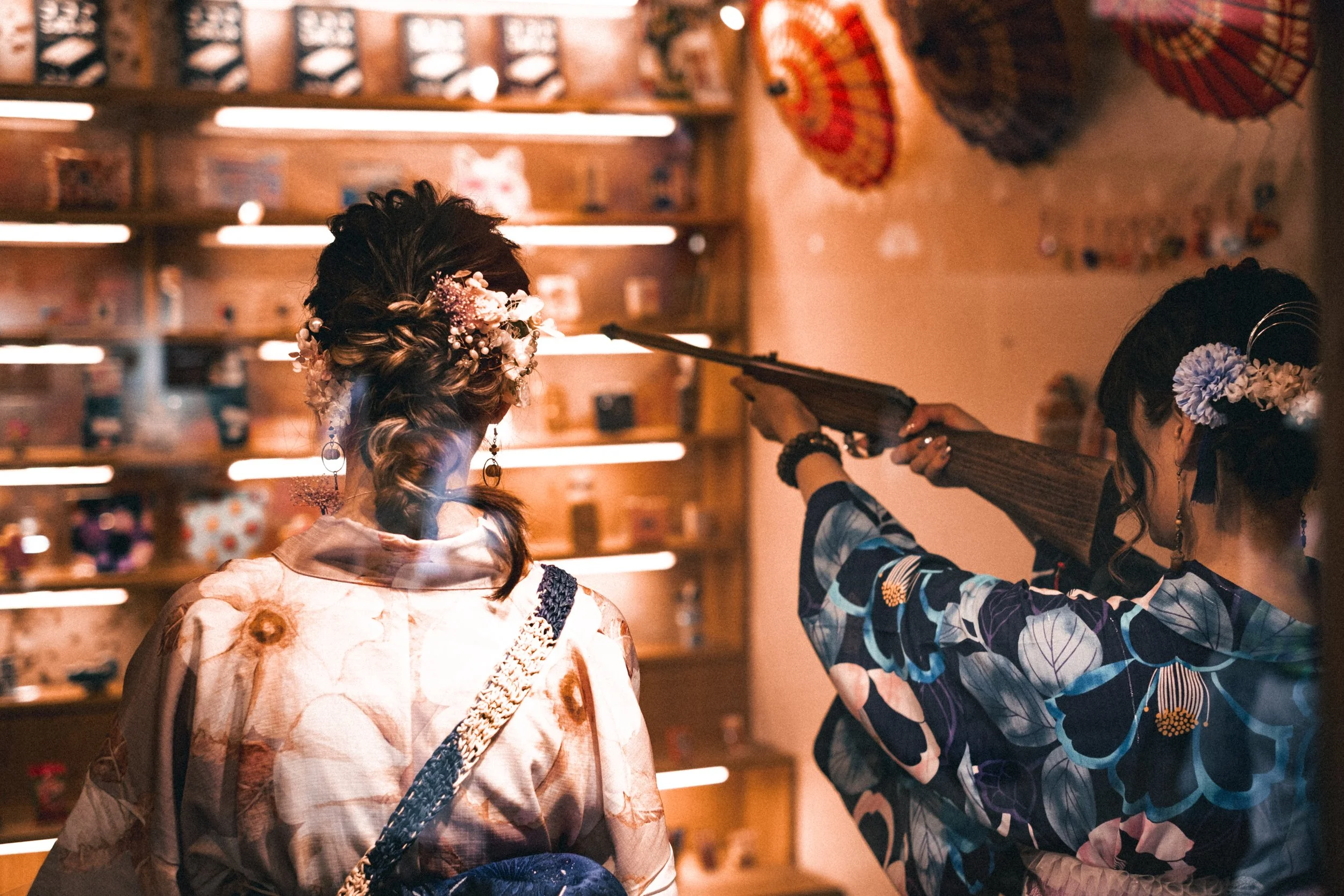 Deux femmes en yukatas à fleurs, dont l’une porte des accessoires floraux dans les cheveux, dans une boutique ou une salle de jeux. L’une vise une vitrine murale remplie de bonbons avec un fusil, de dos à la caméra.