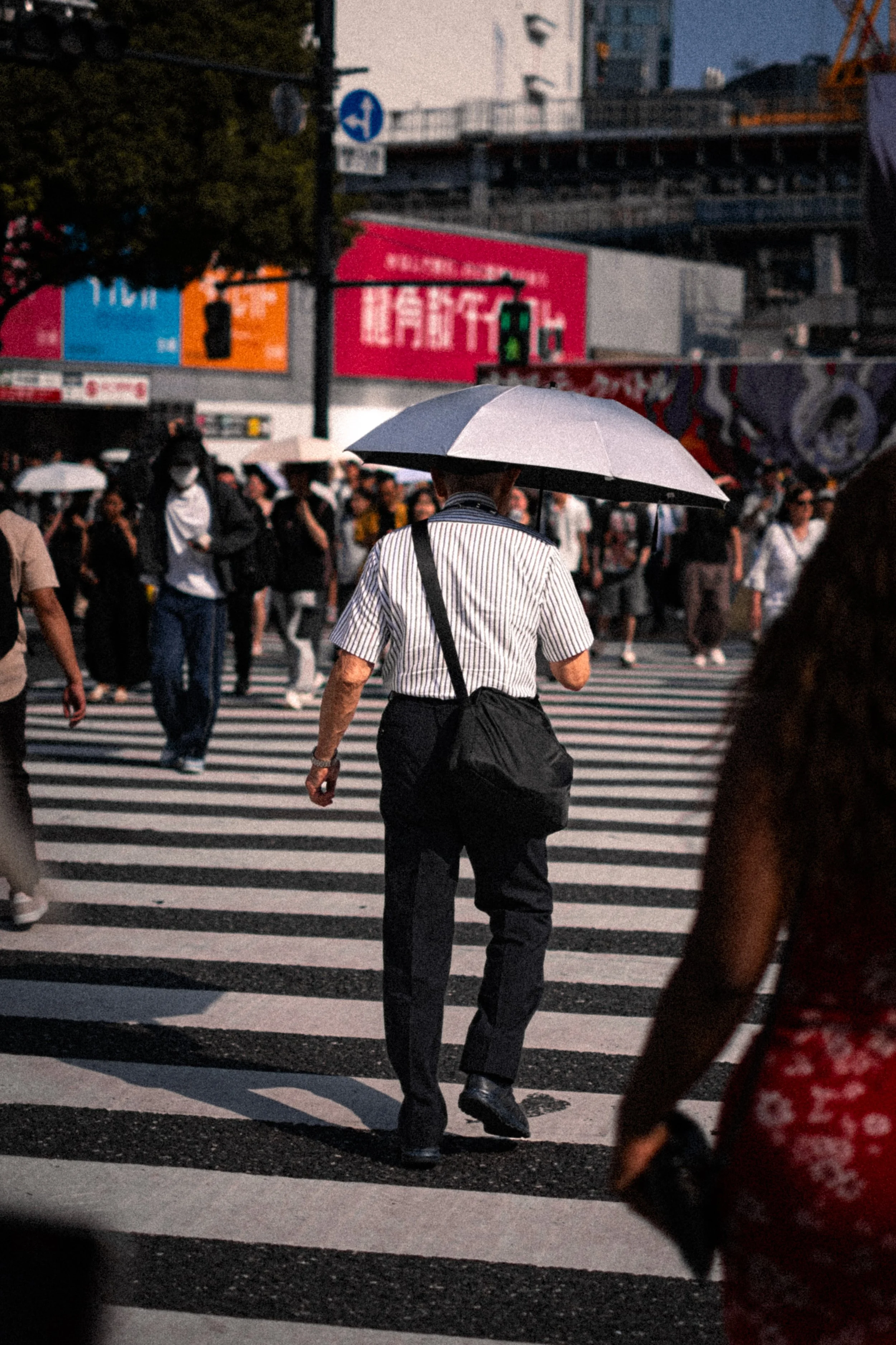 Personne tenant un parapluie gris traversant un passage piéton animé à Shibuya, Tokyo, entourée de nombreux passants et de publicités colorées en arrière-plan.