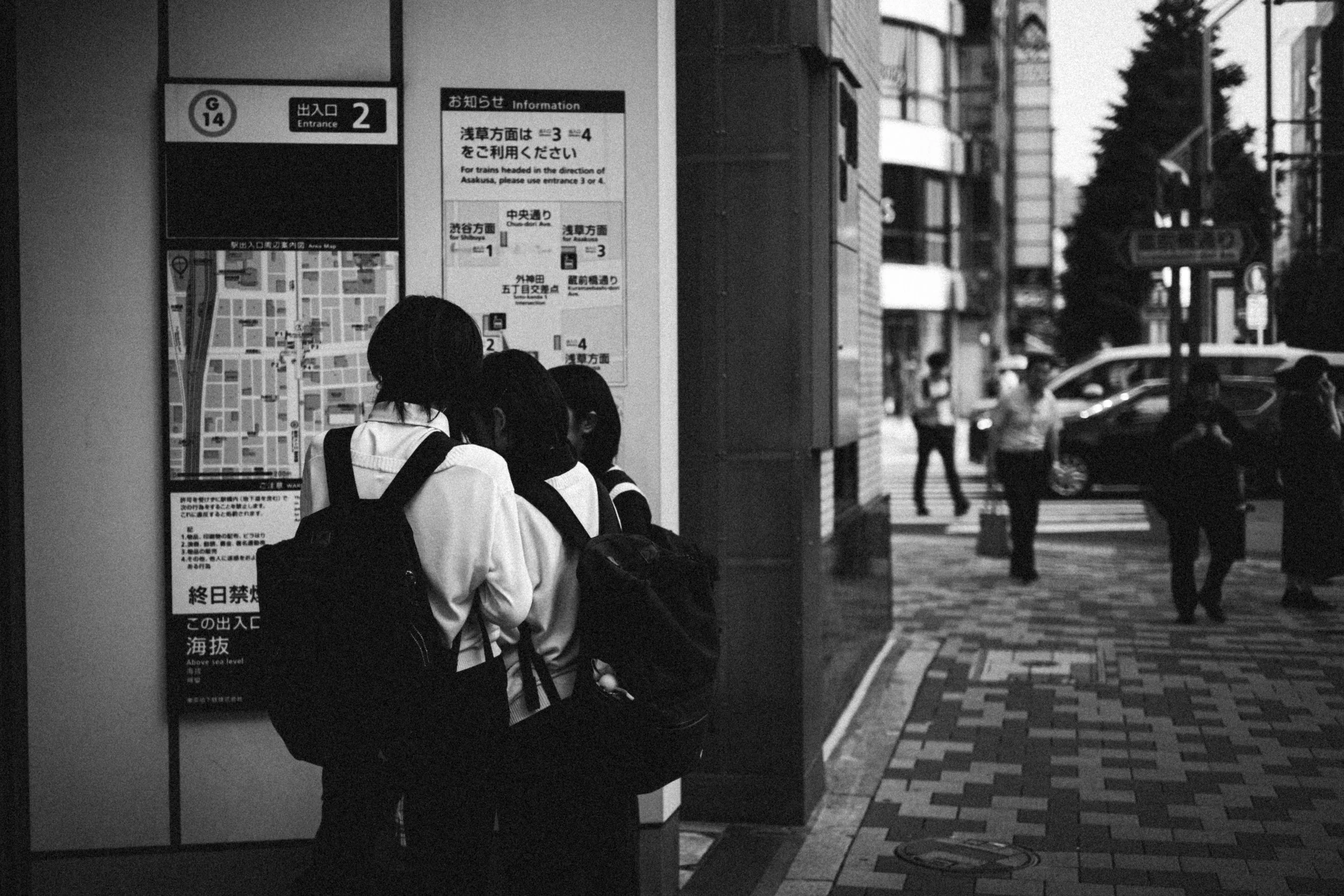 Groupe de collégiennes en uniforme avec sacs à dos, rassemblées autour d’un plan et d’un panneau d’information devant un bâtiment en ville, avec piétons et voitures en arrière-plan.
