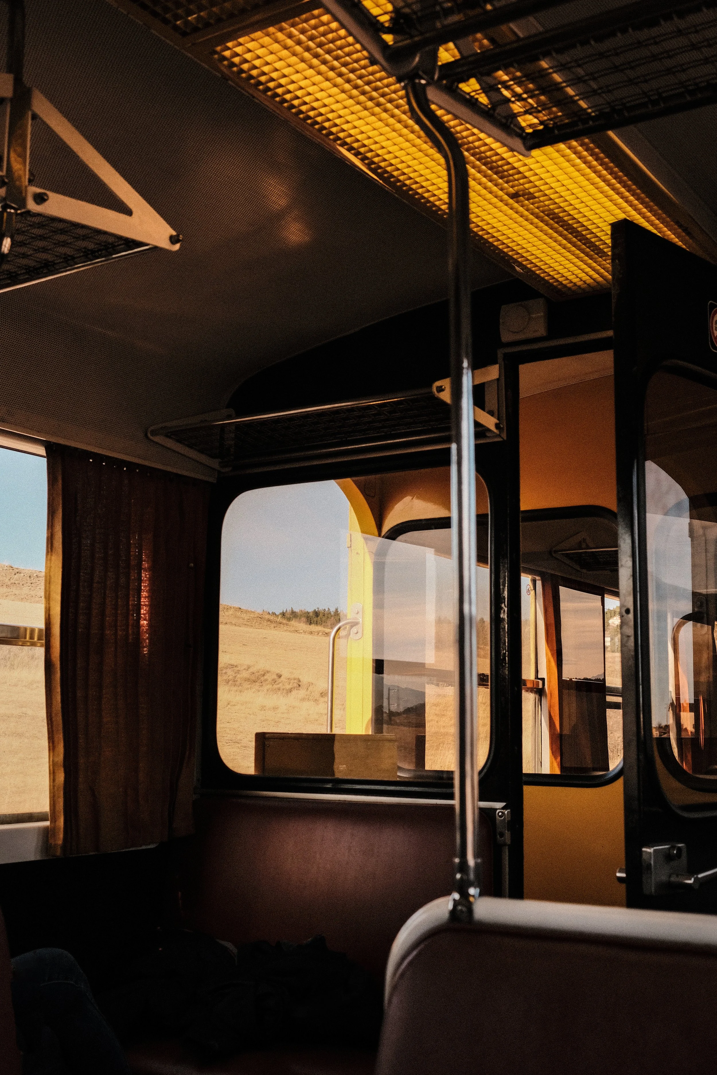 Intérieur d’un train avec grandes fenêtres avant donnant sur un paysage rural de collines et de champs au coucher du soleil, rideaux marron et porte-bagages au plafond.