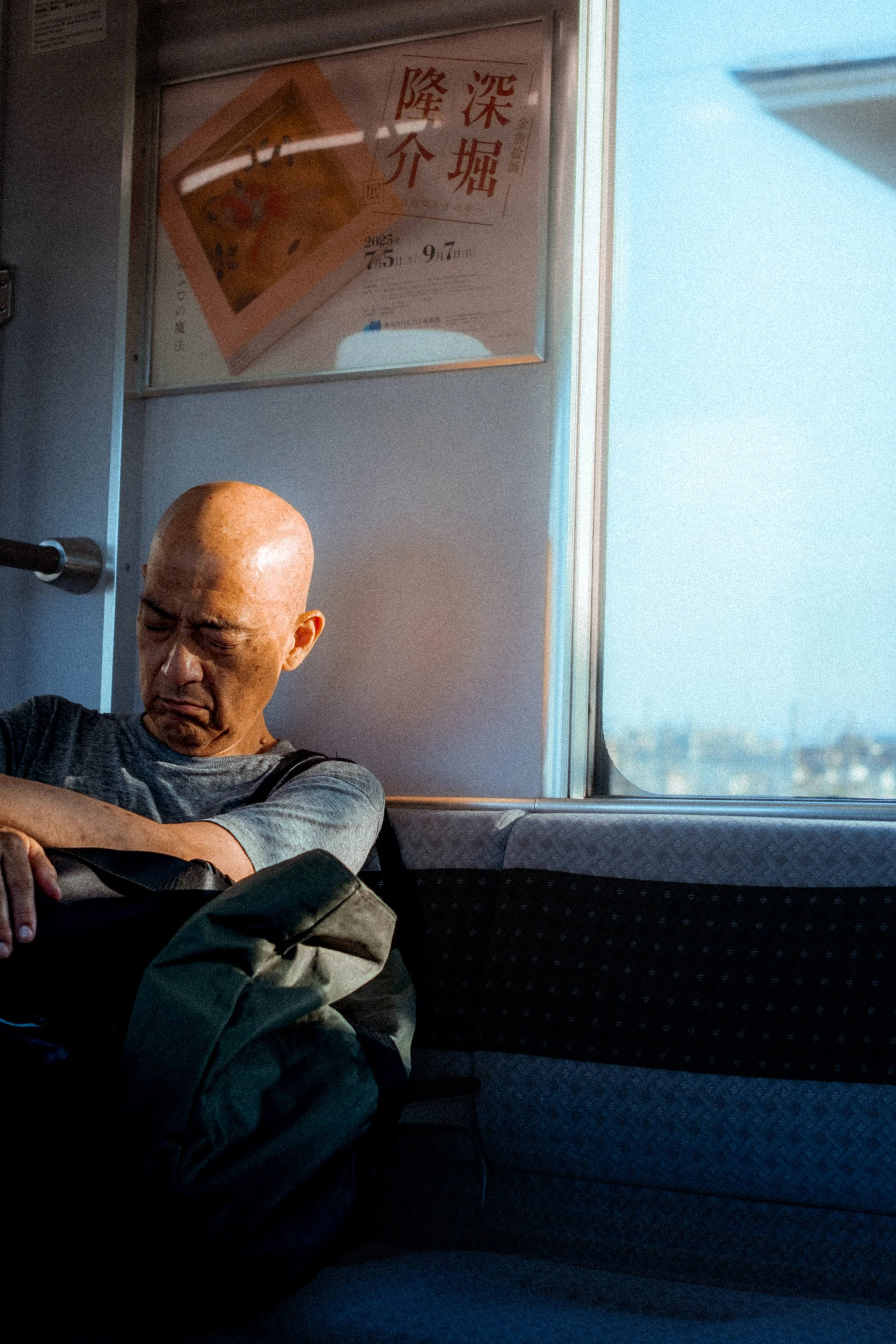 Homme chauve assis dans un train, endormi, sac à dos sur les genoux. À l’arrière-plan, une fenêtre montre un paysage urbain flou et une affiche avec des caractères japonais.