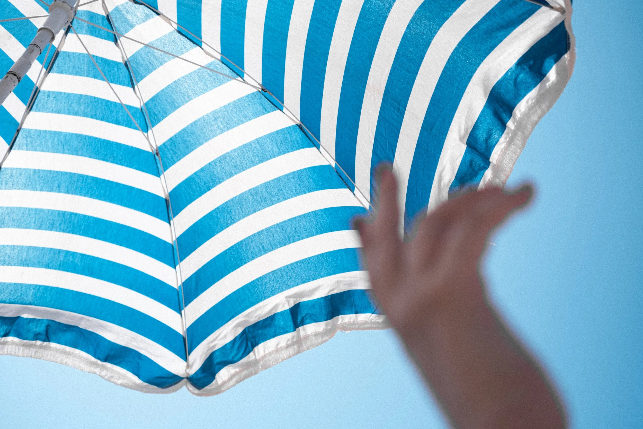 Gros plan sur un parasol de plage à rayures bleues et blanches sous un ciel bleu clair, avec une main tendue vers lui.