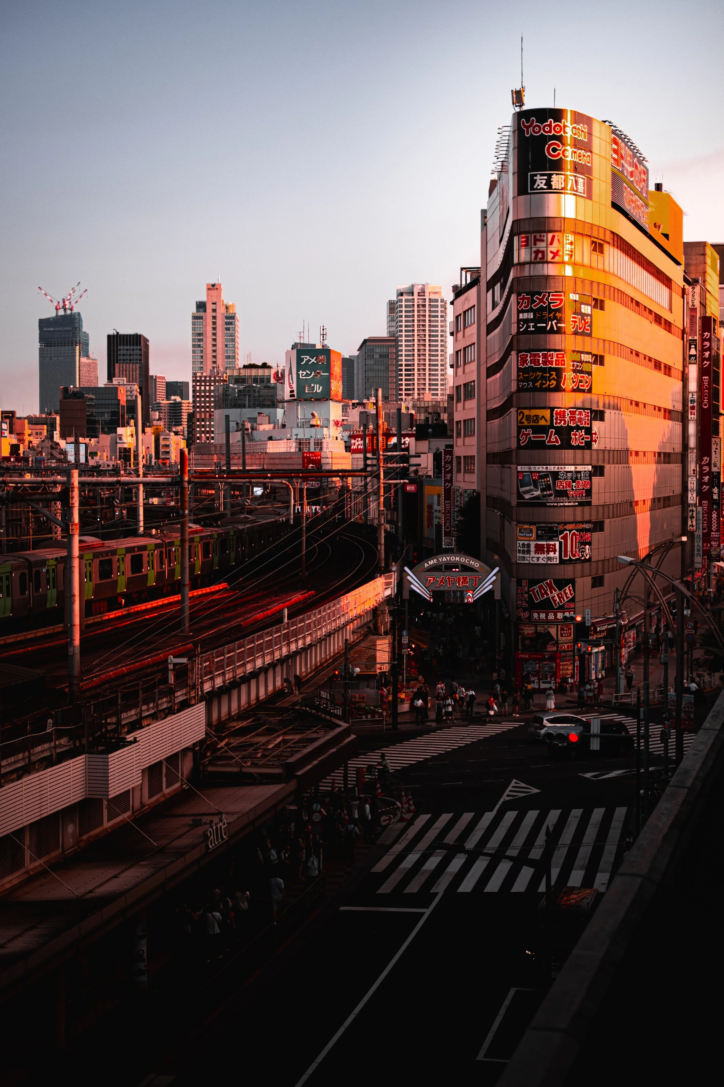 Paysage urbain de Tokyo au coucher du soleil avec de hauts immeubles, enseignes néon en japonais et un train circulant sur des voies surélevées.