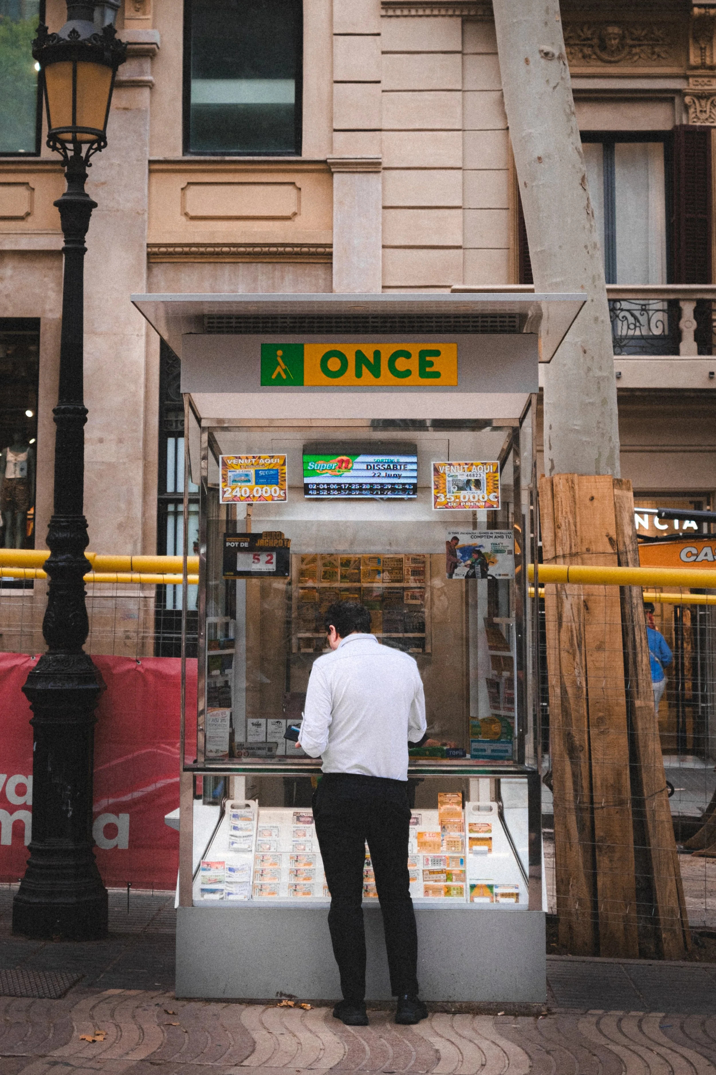 
Homme en chemise blanche et pantalon noir debout à un kiosque de loterie sur un trottoir en ville, avec un lampadaire et un immeuble en arrière-plan.