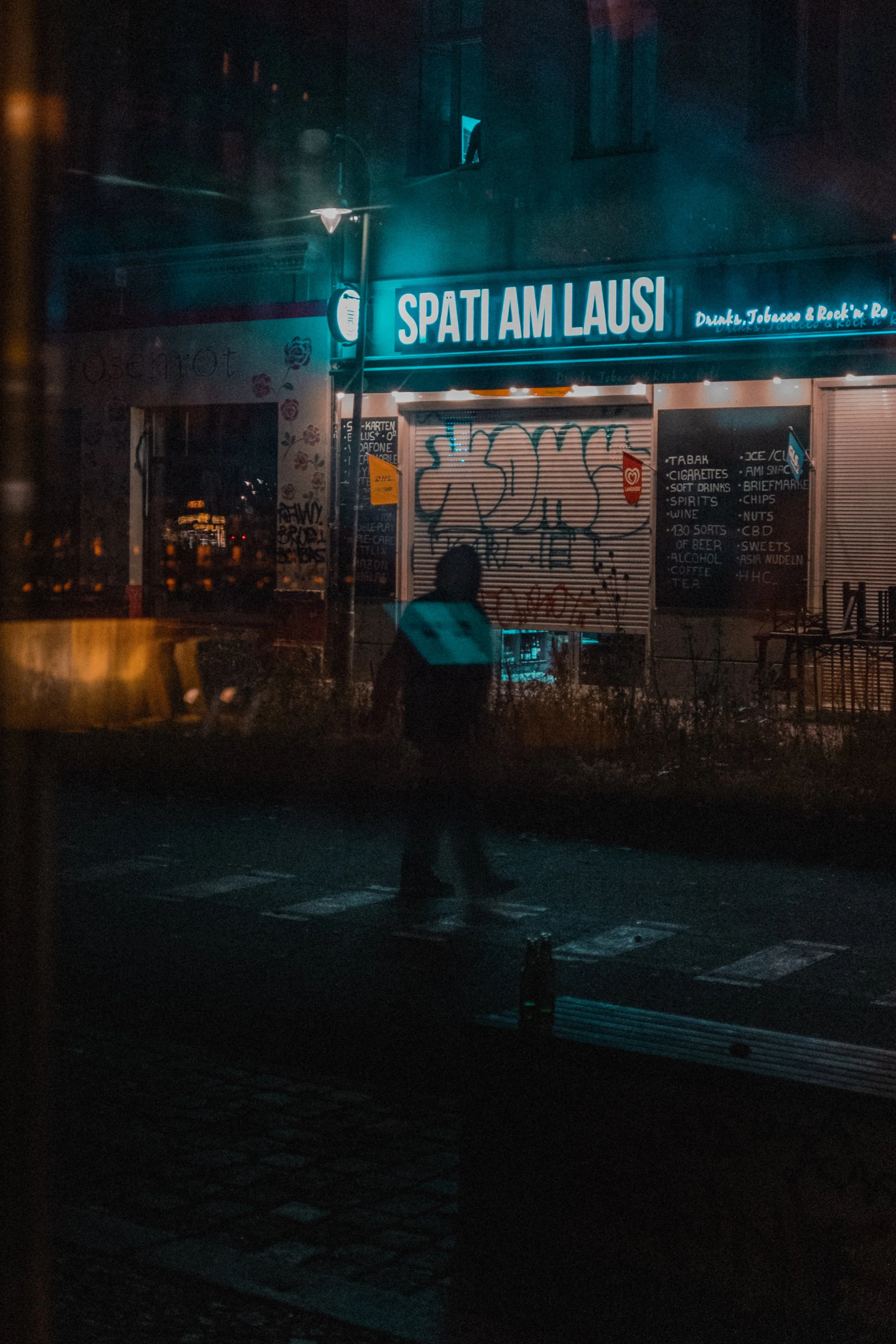 
Scène nocturne avec une personne passant devant une boutique fermée couverte de graffitis, enseigne néon bleue "SPAT AM LAUSI", panneau "Drinks, Tobacco & Rock in R", menu sur ardoise listant cigarettes, boissons et alcool, reflets de lumières