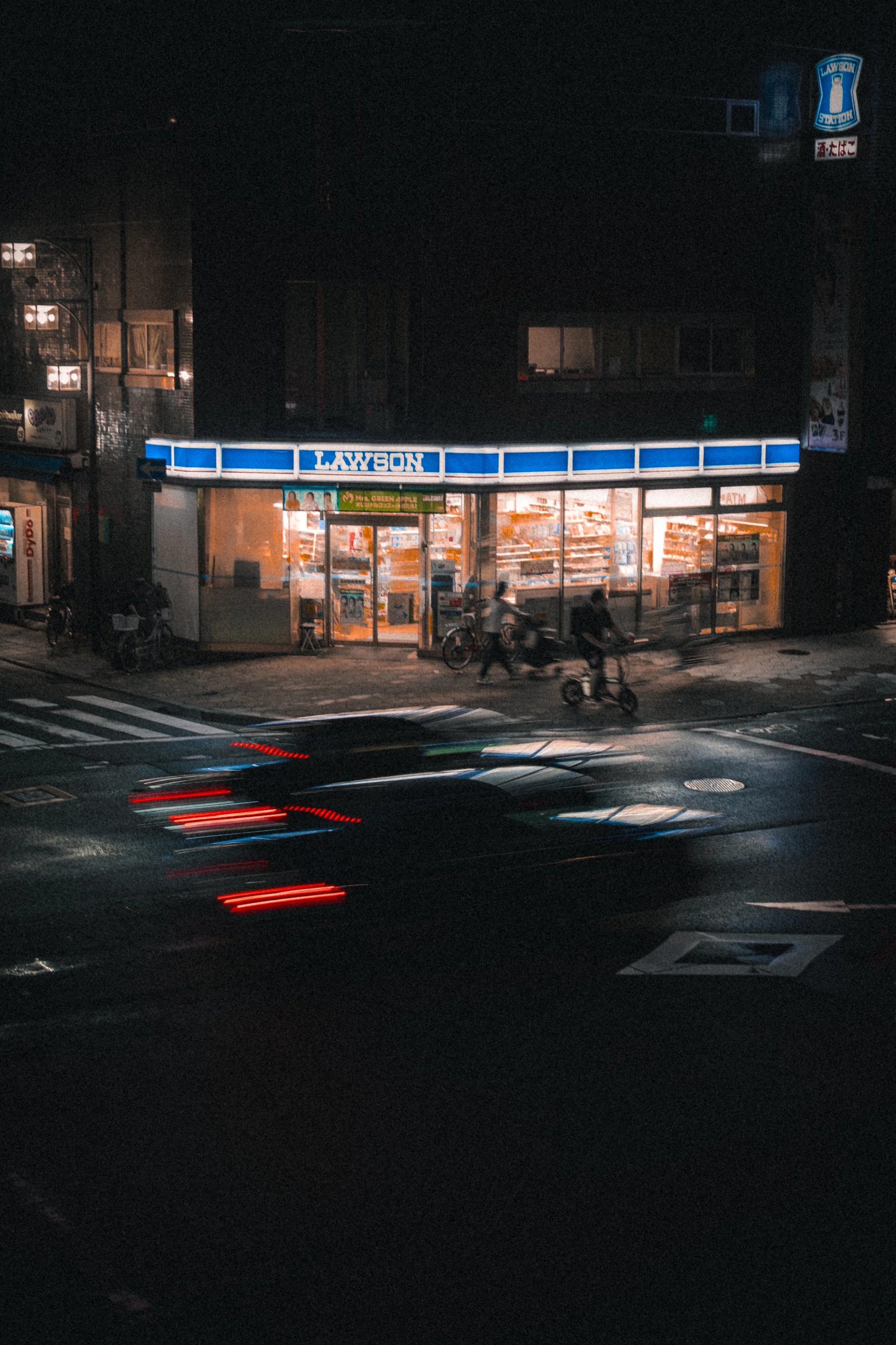 Supérette Lawson à Osaka vivement éclairée la nuit, avec des piétons et des vélos à l’extérieur, et des phares de voiture créant des traînées lumineuses sur la rue sombre au premier plan.
