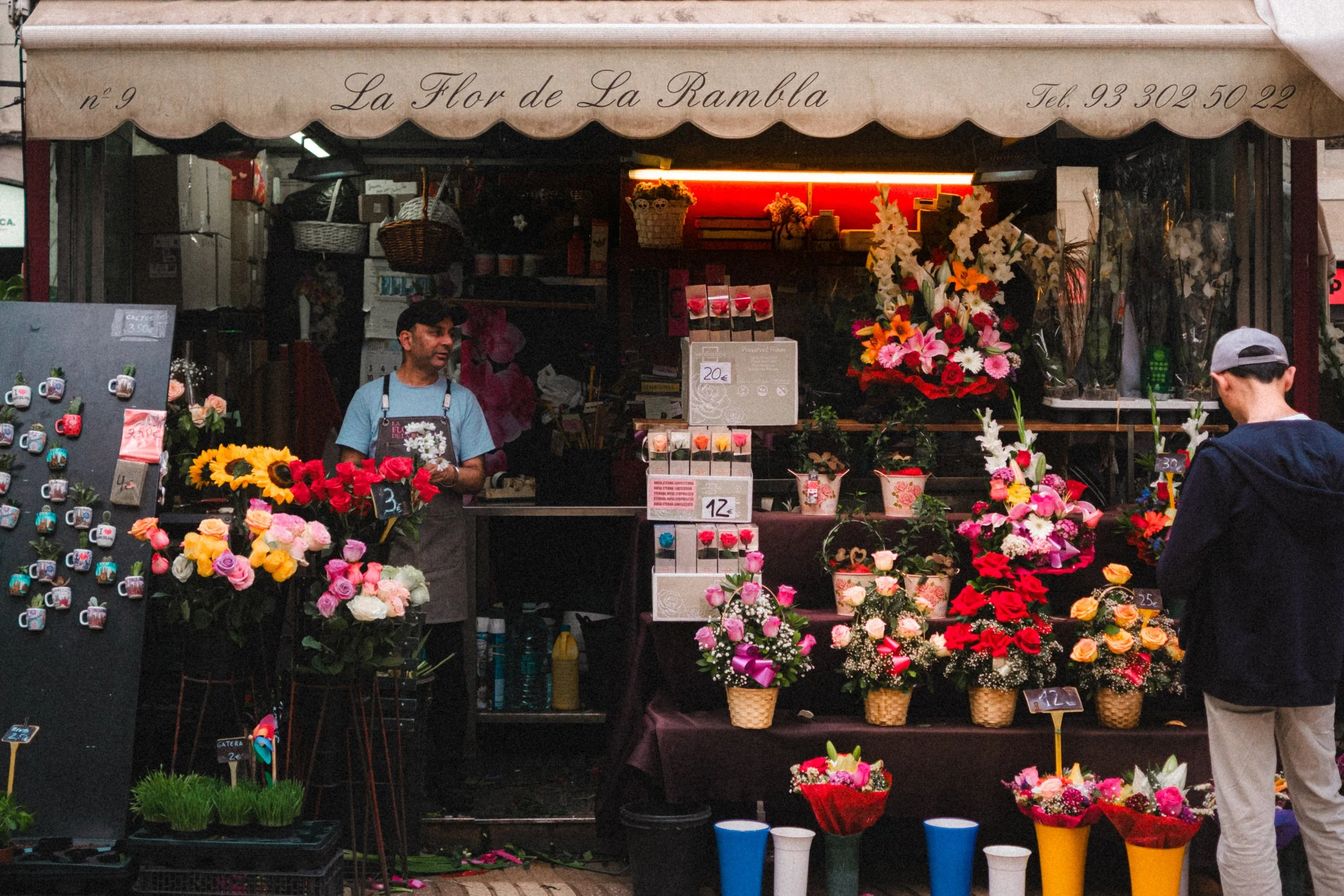 Stand de fleuriste avec diverses fleurs colorées, dont des roses et des lys, exposées sur des étagères et dans des paniers. Un vendeur et un client parcourent les arrangements floraux