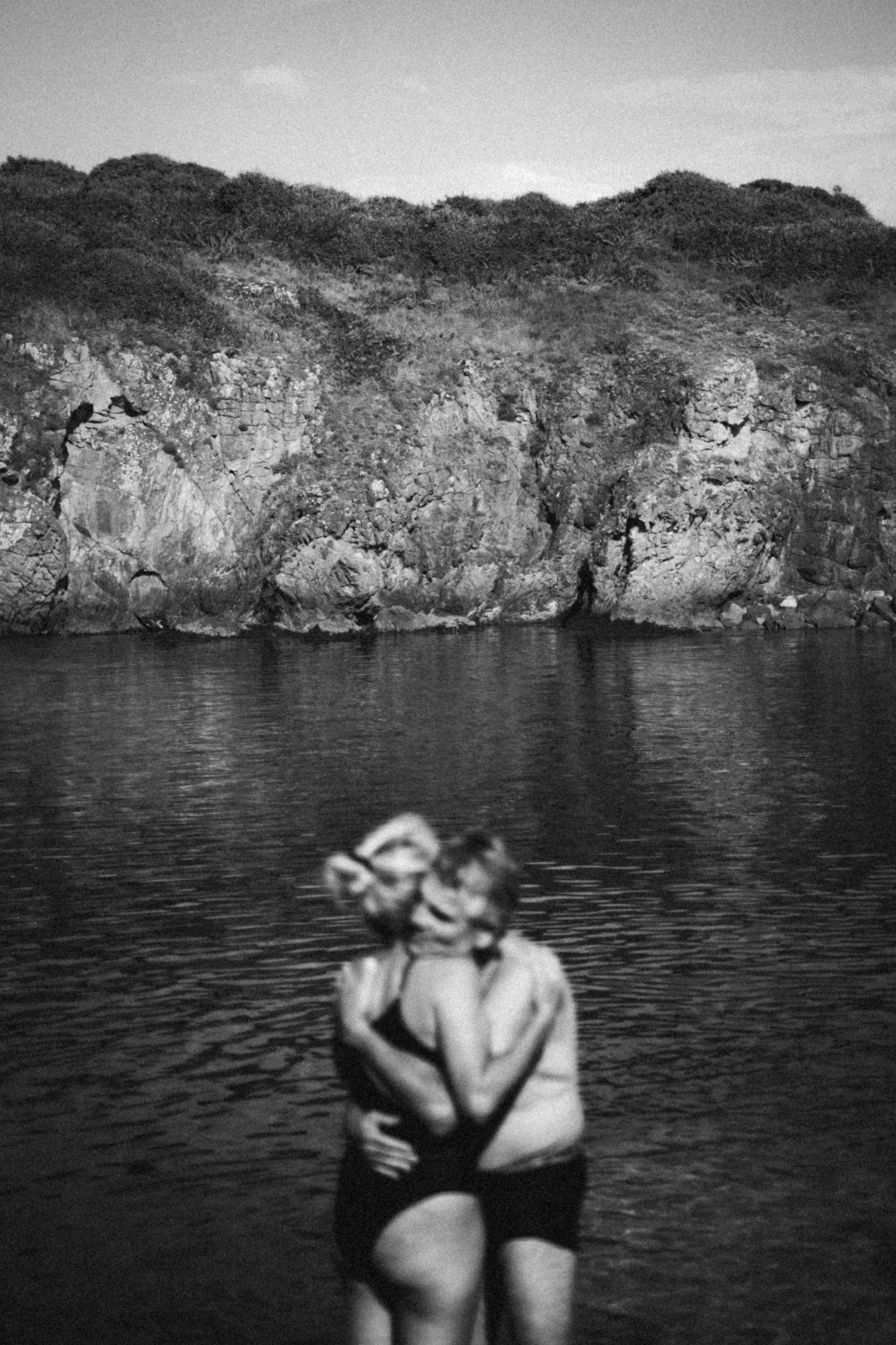 Deux individus s’enlaçant et s’embrassant dans l’eau près d’un rivage rocheux, avec des buissons et une colline en arrière-plan.

