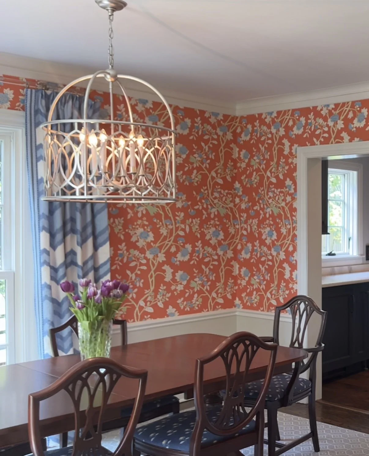 Dining room with orange floral wallpaper, a wooden table with four chairs, a vase of purple tulips, a chandelier, and a window with blue and white patterned curtains.