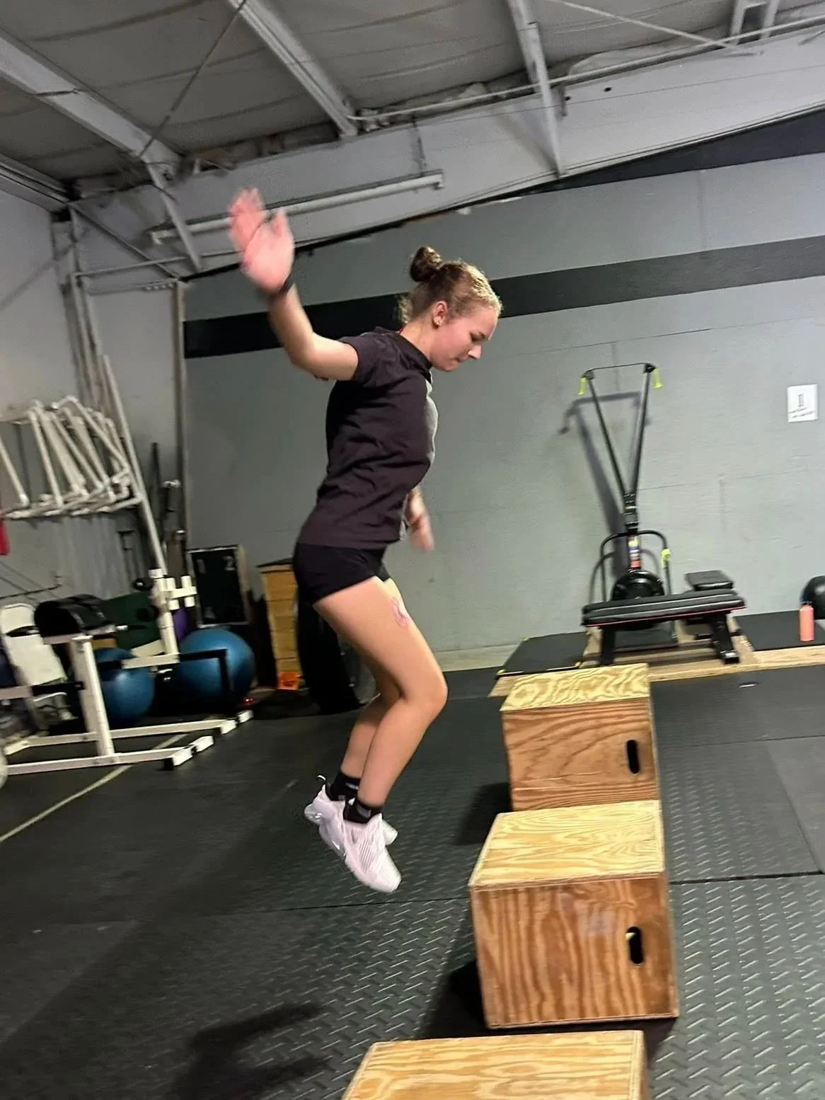 A woman jumping onto a wooden plyometric box at a gym. She is mid-air with arms raised, wearing a black t-shirt, black shorts, and white athletic shoes.