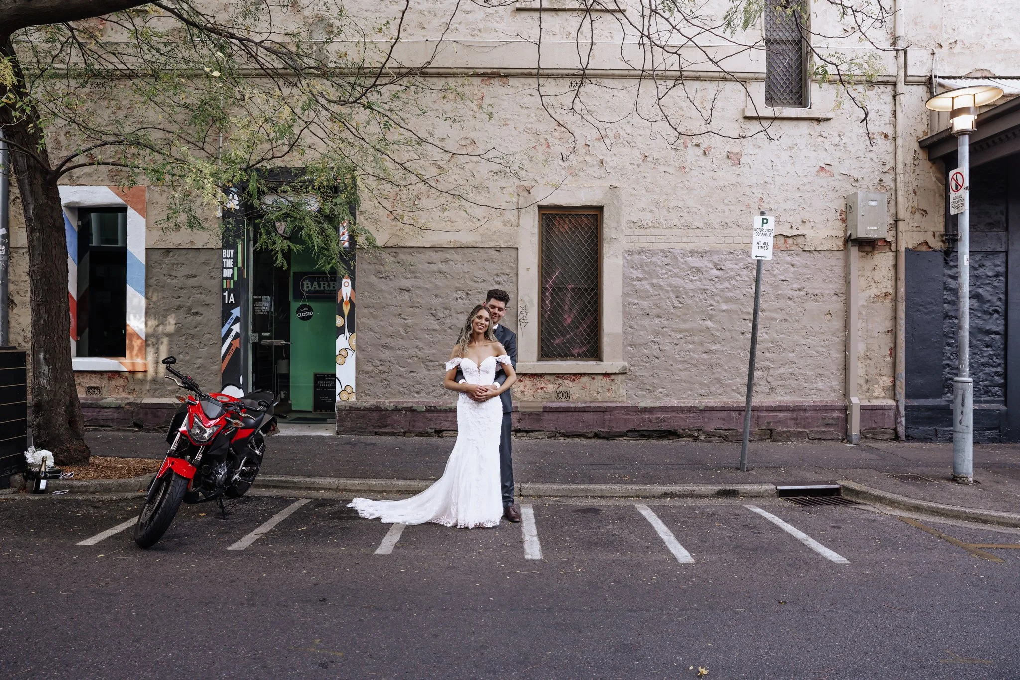 A newlywed couple stands on an empty city street, with the bride in a white wedding gown and the groom in a suit, embracing each other in front of a weathered brick building. A red motorcycle is parked on the street nearby.