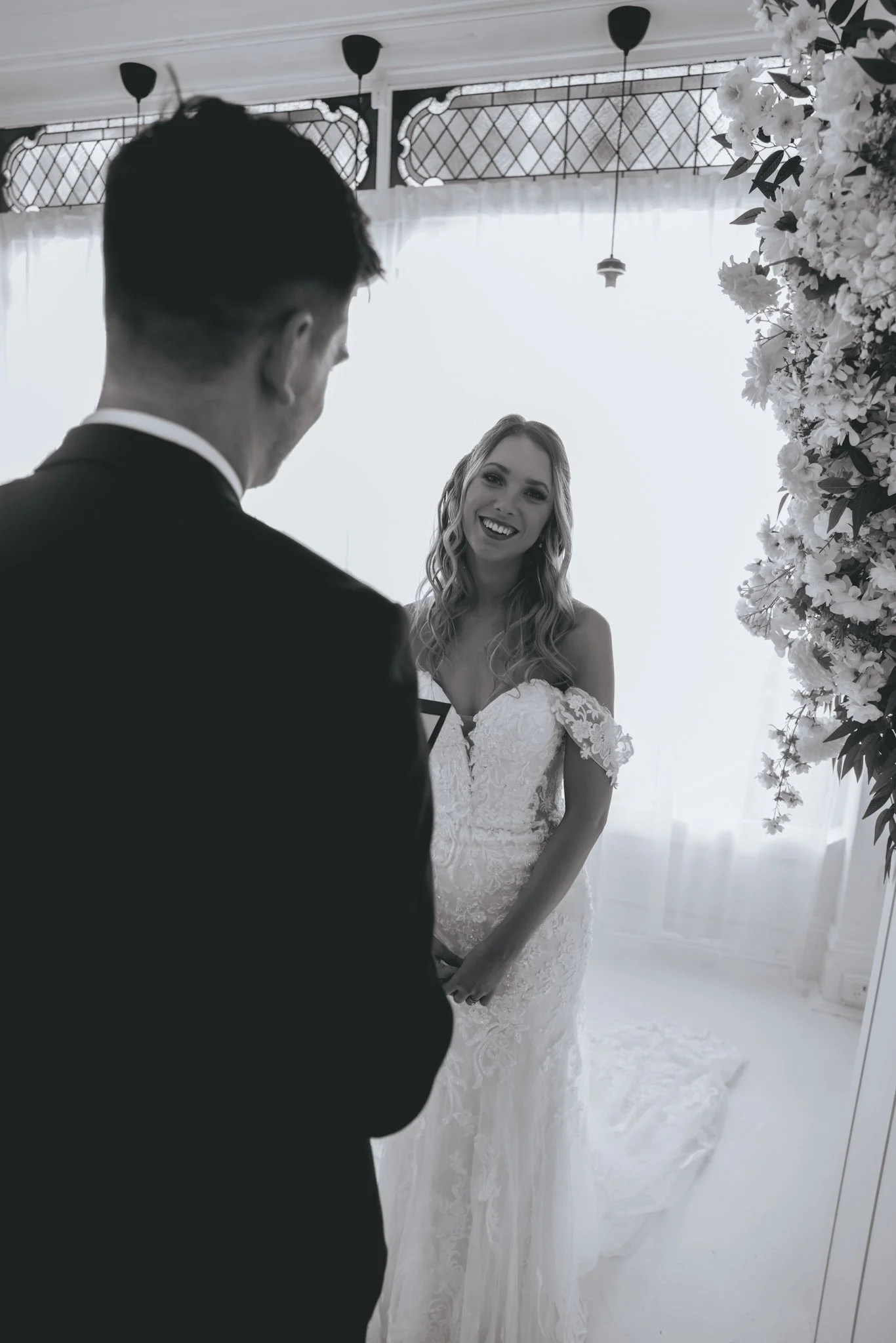 A bride in a white wedding gown smiling at a groom in a black suit during a wedding ceremony in a bright room with floral decor.
