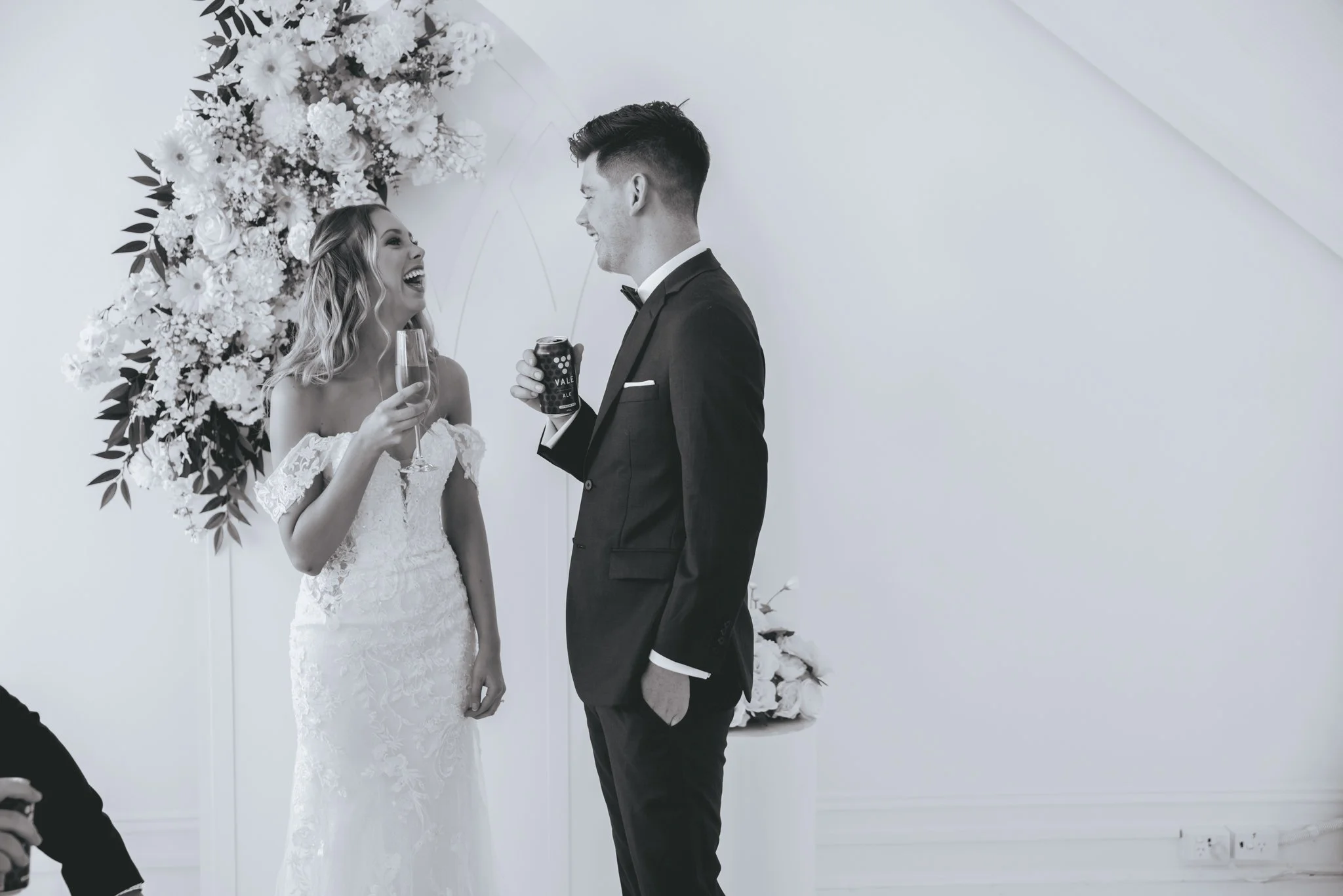 A bride and groom happily talking during their wedding, with the bride holding a glass of champagne and the groom holding a drink can, standing near a floral wedding backdrop.