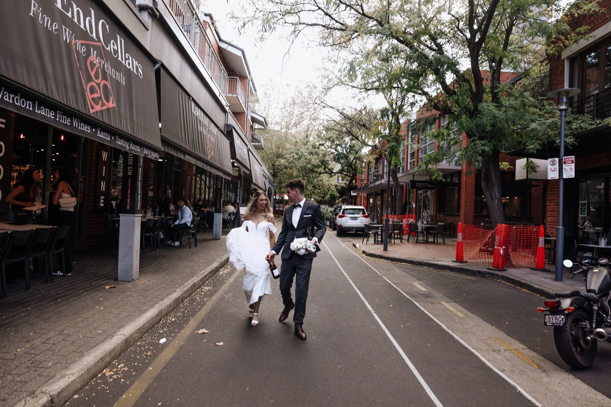 A newlywed couple walking down a city street, holding hands and smiling. The bride is wearing a white wedding dress and the groom is in a black suit. The street is lined with cafes and shops, some people sitting outside.
