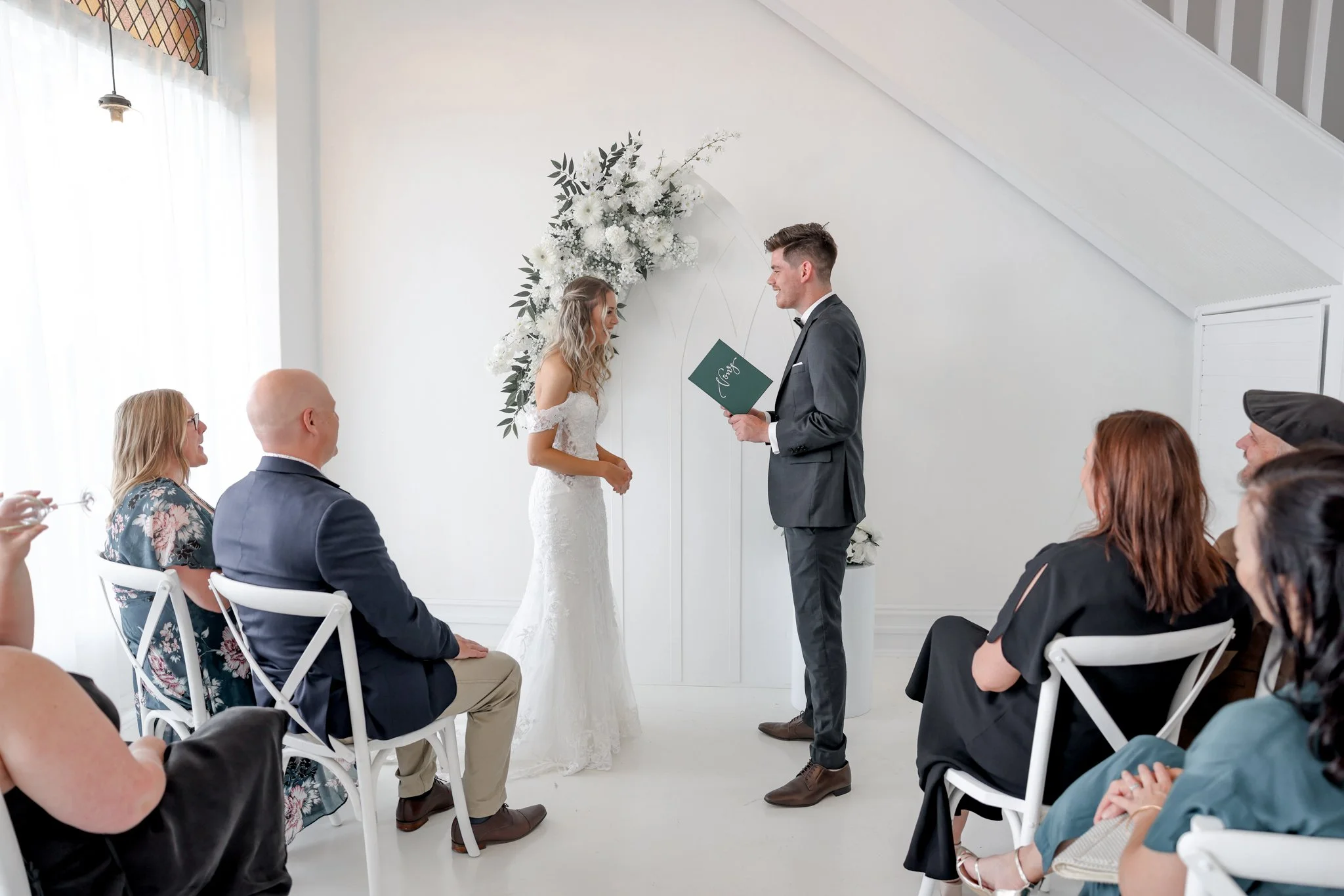 A couple getting married during a wedding ceremony in an indoor setting with white walls and a high ceiling. The bride, in a lace wedding gown, and the groom, in a gray suit, stand facing each other. The groom holds an open book or folder. Behind the