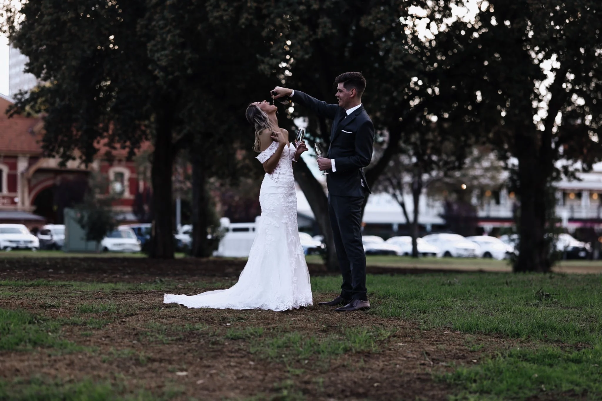 A bride and groom celebrating outdoors, the groom is pouring champagne into the bride's mouth.