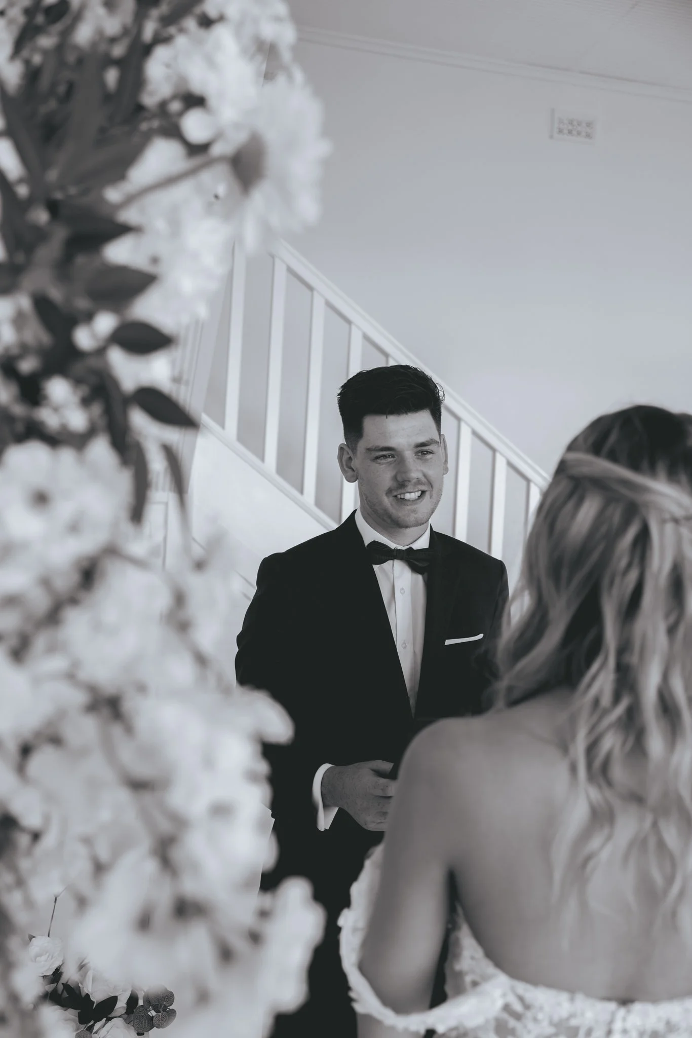 A groom in a tuxedo holding hands with a bride in a wedding dress during their wedding ceremony, with a floral arrangement in the foreground.
