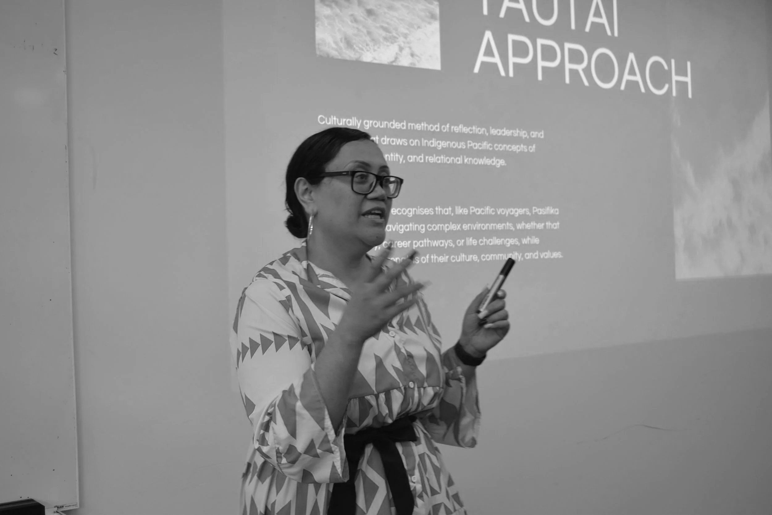 A woman wearing glasses and a patterned shirt during a presentation, gesturing with her hands and holding a phone, with a projected screen and text in the background.