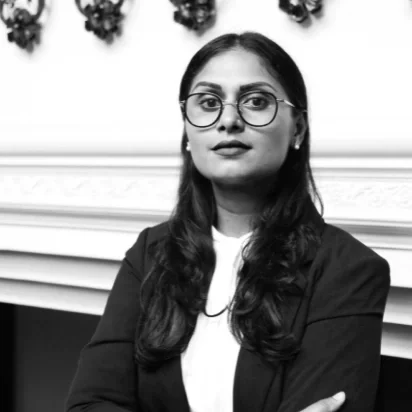 A woman with glasses and long dark hair, wearing a blazer and white top, sitting with arms crossed in front of a decorated white fireplace mantle in black and white.