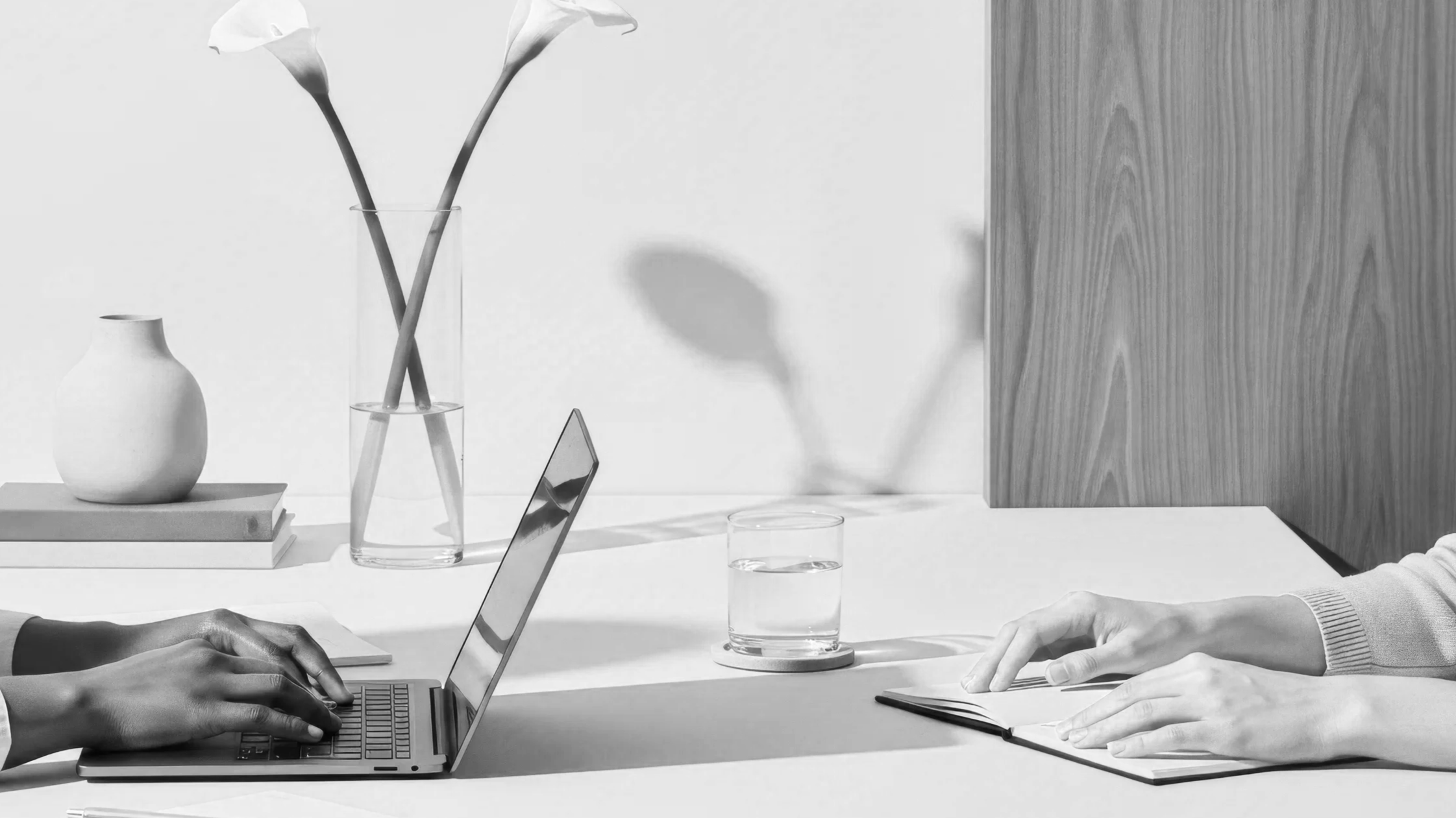 Two people in business attire working at a white table with laptops, notebooks, a glass of water, and decorative vases on the table.