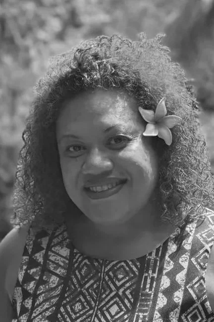 Woman with curly hair and a flower in her hair smiling at the camera, outdoor background.