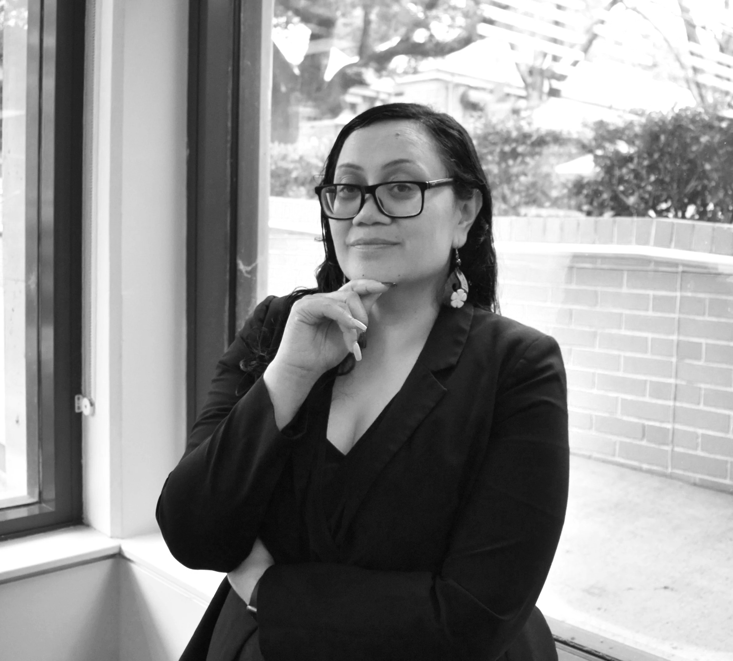 A woman with glasses, earrings, and dark hair, sitting by a window, with her hand near her chin, looking slightly off to the side.
