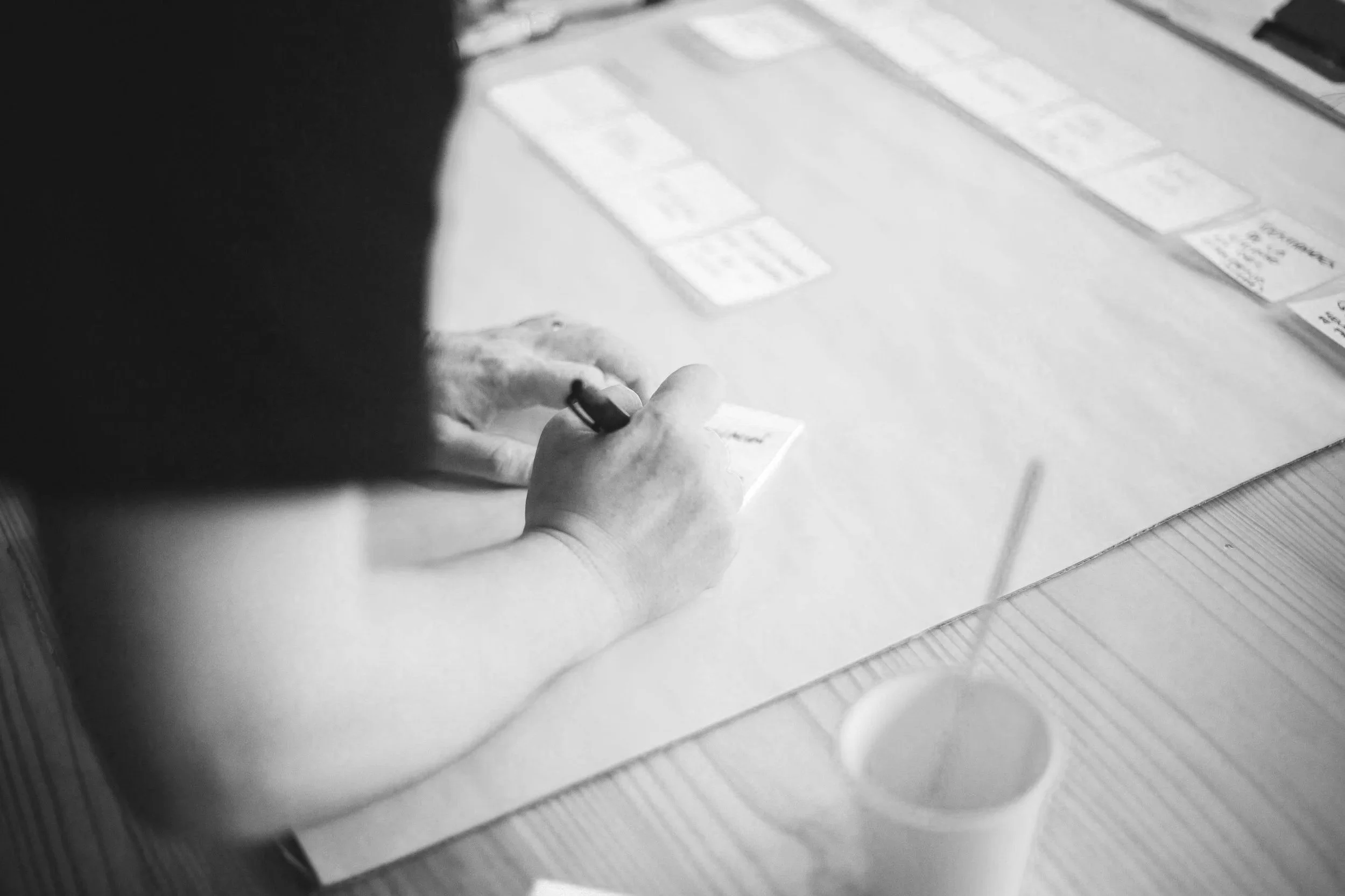 Black and white photo showing a person's hands writing on a notepad or paper, placed on a table behind other sheets of paper or notes.