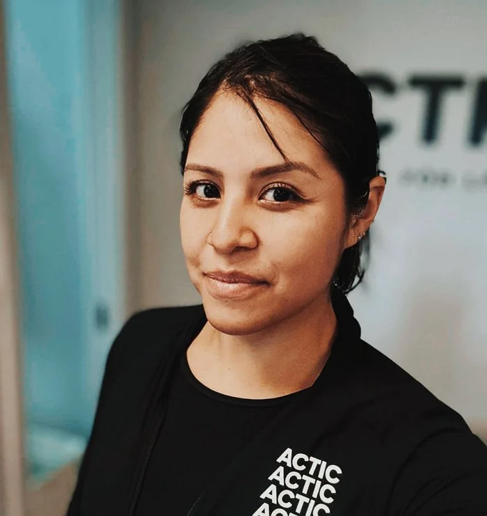 Young woman with dark hair tied back, wearing a black shirt with the words 'ACTIC' repeated on it, standing indoors in front of a blurred background.