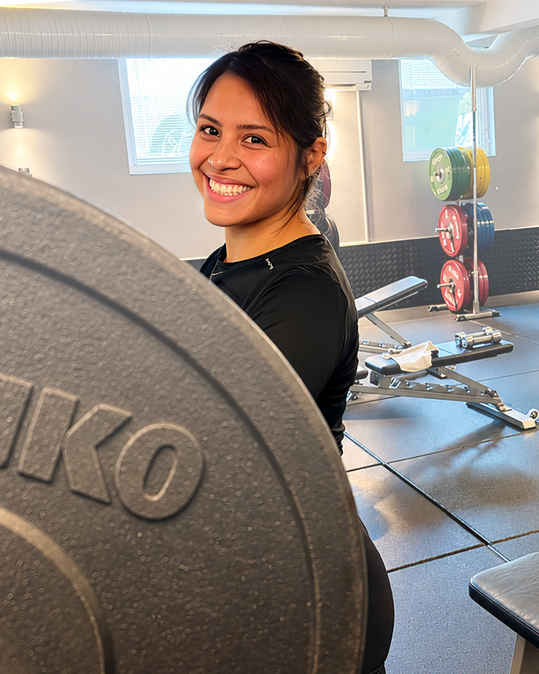 Glad kvinna i träningskläder i gymmet, bakom viktplattor och träningsutrustning.