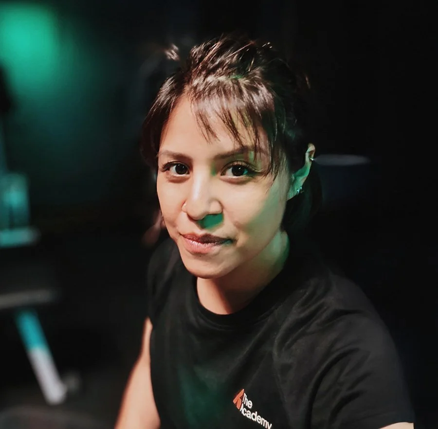 A woman with short dark hair and light skin, wearing a black shirt, is taking a selfie in a dimly lit environment with blue and green lighting.