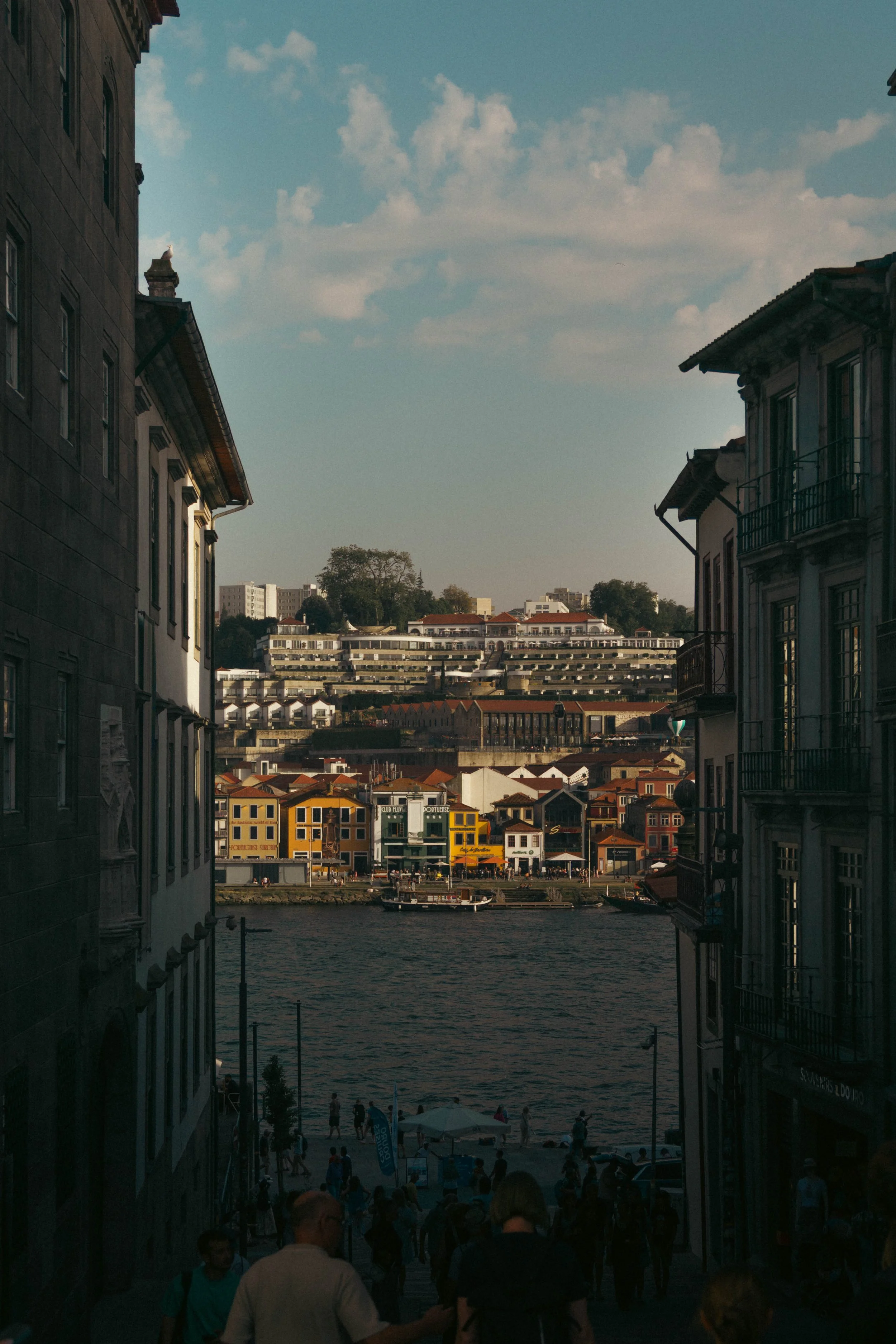 View of a waterfront cityscape with colorful buildings on the riverbank, a boat docked along the shore, and a hillside with residential structures in the background under a partly cloudy sky.