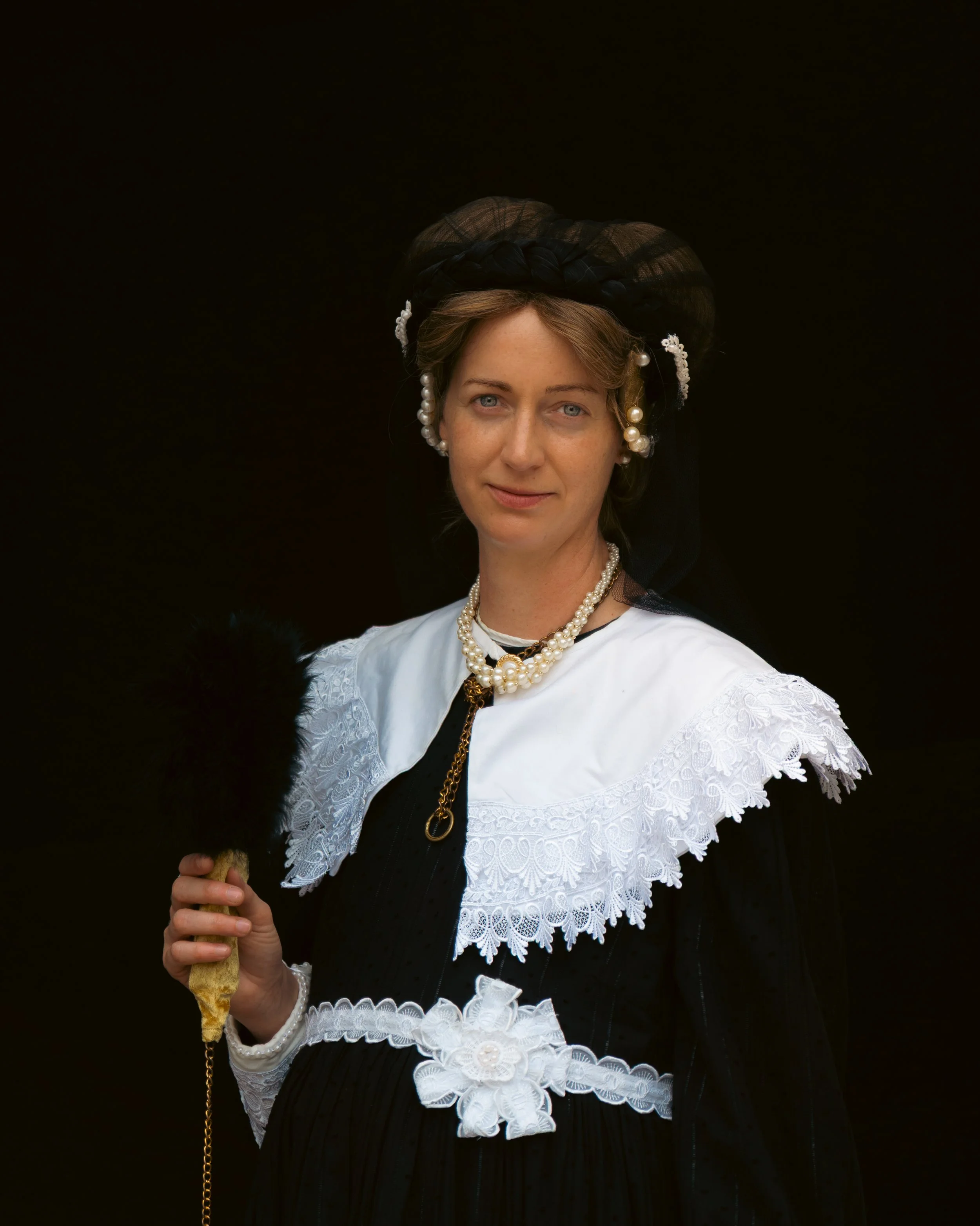 Woman dressed in traditional European clothing with lace details, pearl jewelry, and a black headscarf, holding a feathered fan against a dark background.