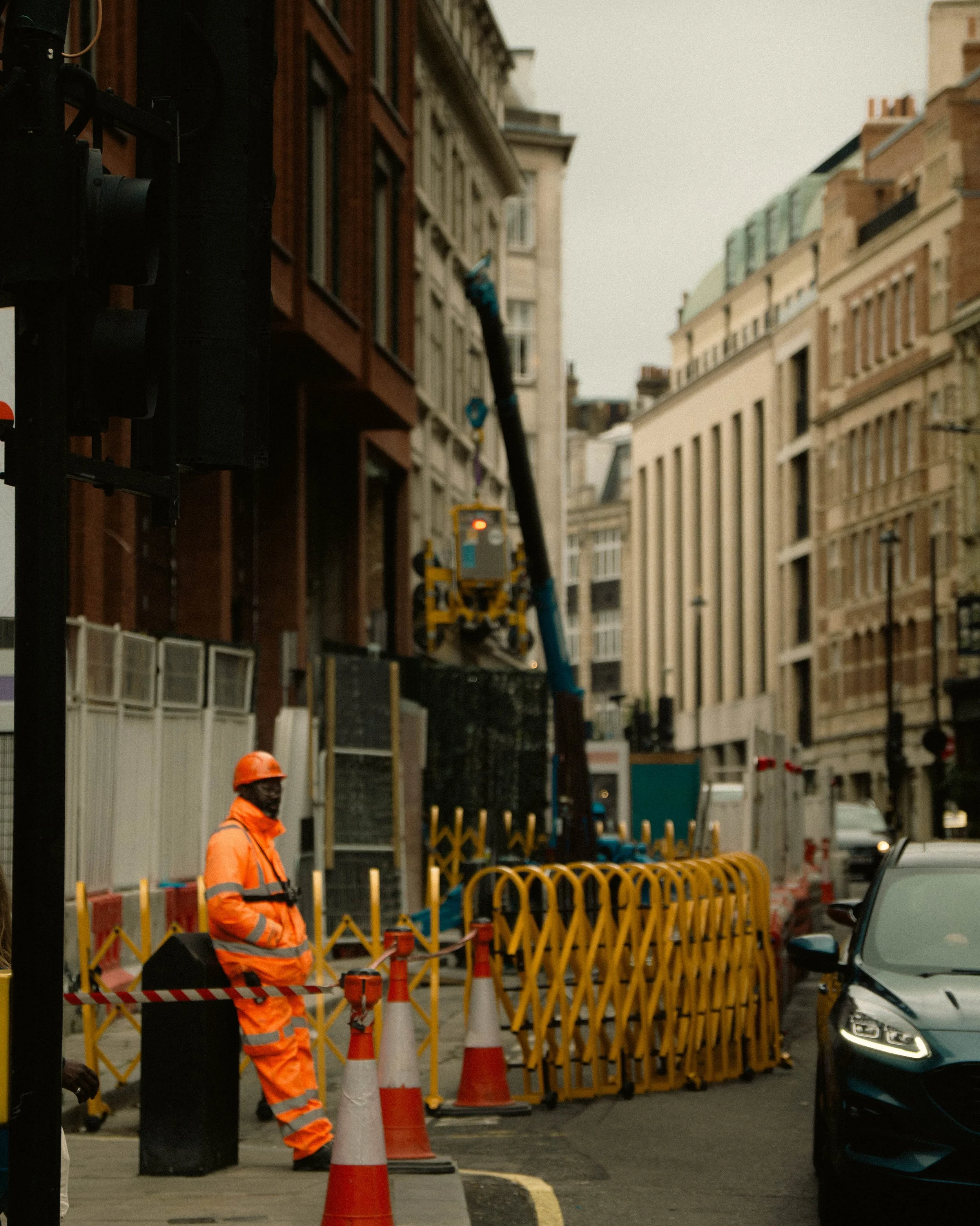 City street construction scene with a worker in orange safety gear, orange and white traffic cones, barriers, and construction equipment, with tall buildings in the background.