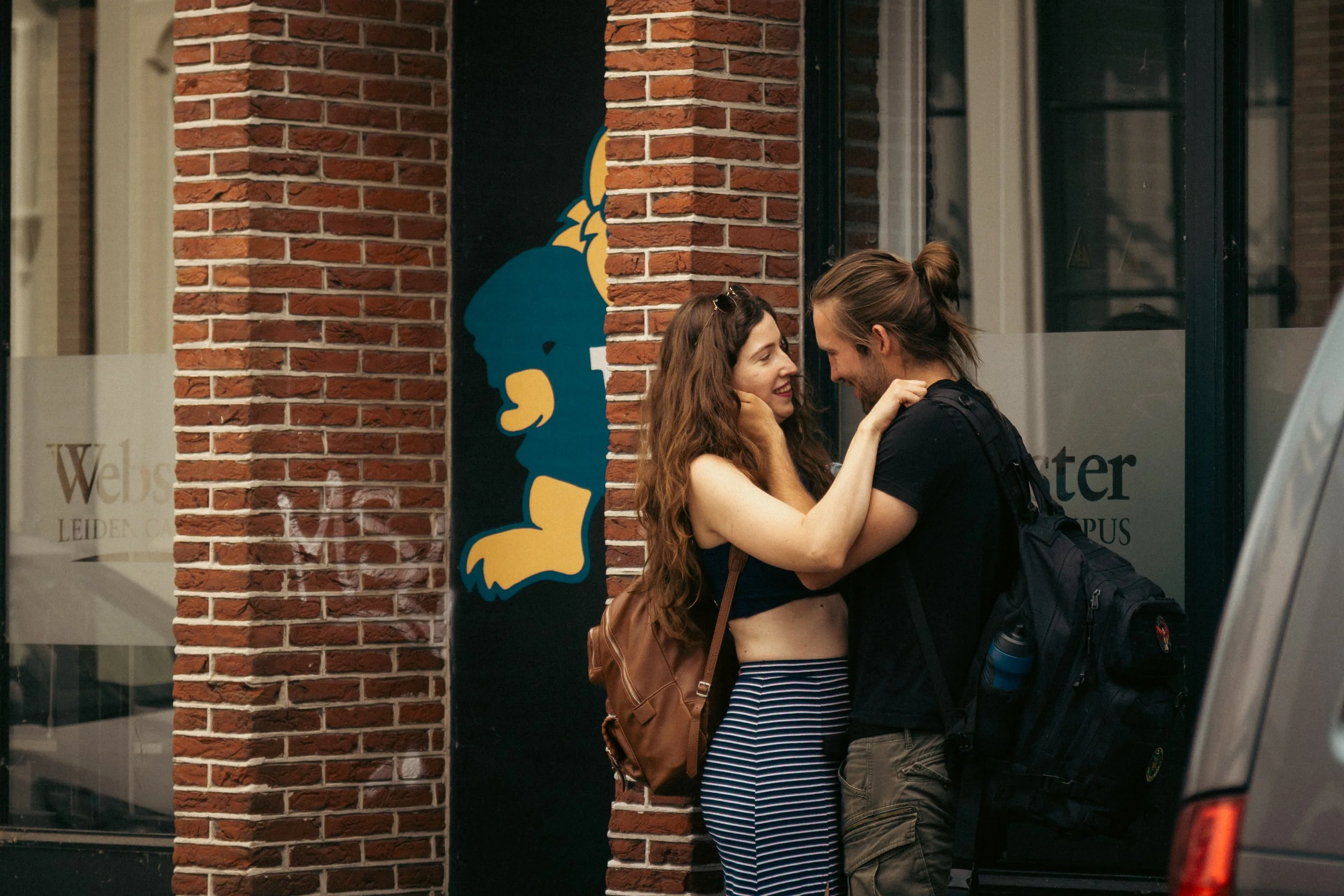 A young couple stands close together outdoors, smiling and touching faces near a brick building with a cartoon lion painted on the window.
