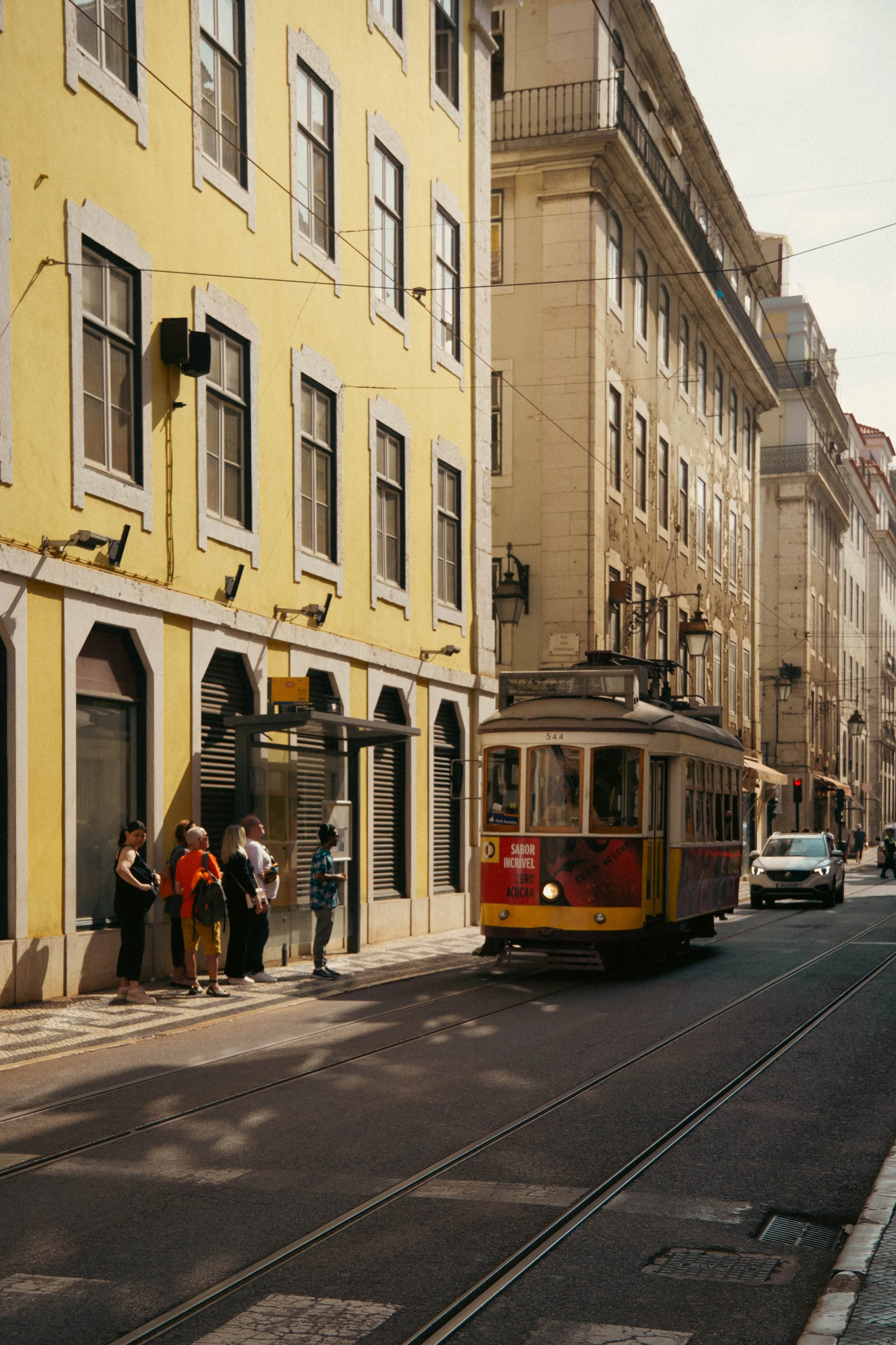 A street scene with a vintage tram approaching a bus stop where several people are waiting, in front of yellow and beige buildings with multiple windows.