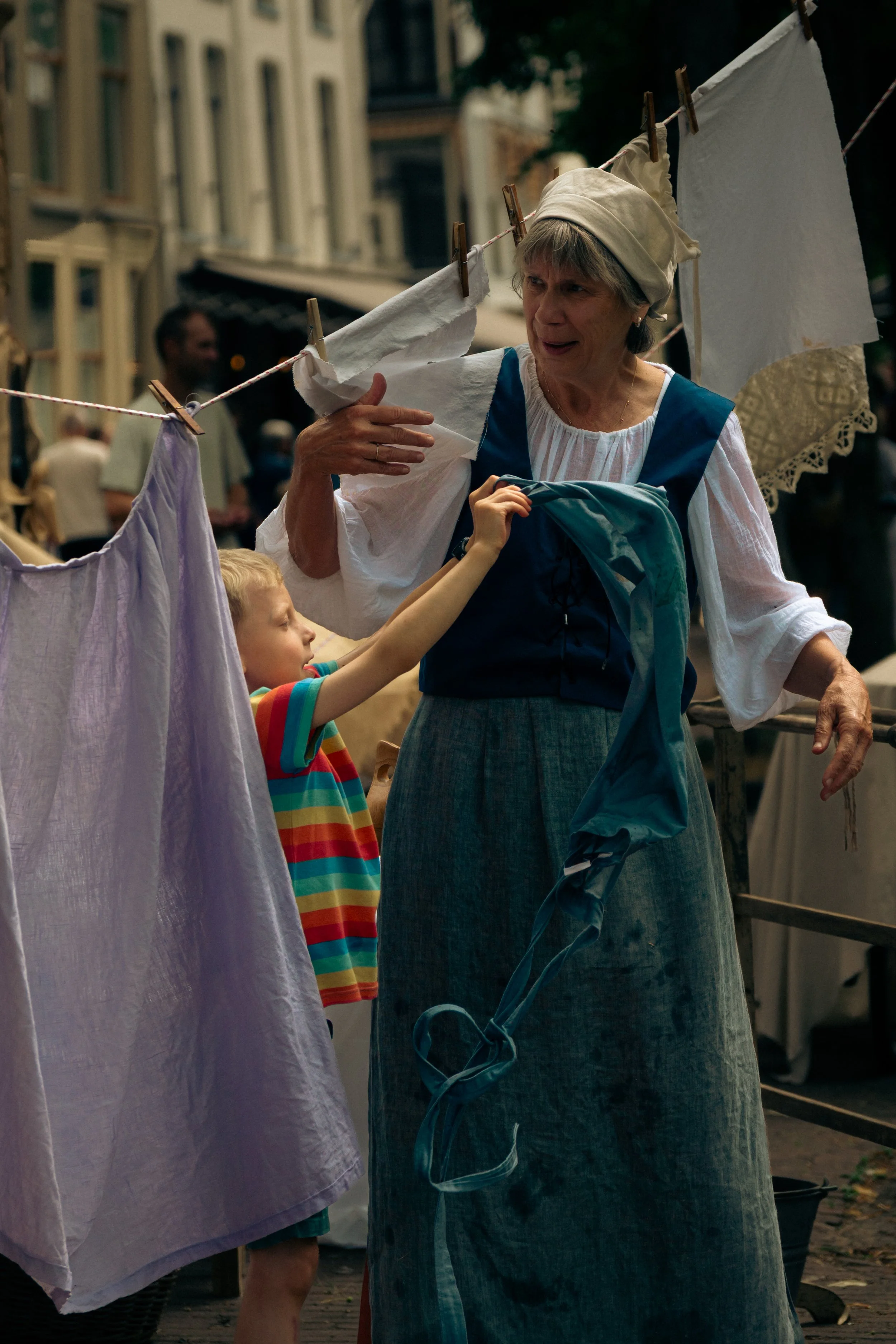 A young boy in a rainbow striped shirt is handing laundry to an older woman in a historical costume, who is standing outdoors among hanging sheets, at what appears to be a street fair or reenactment.