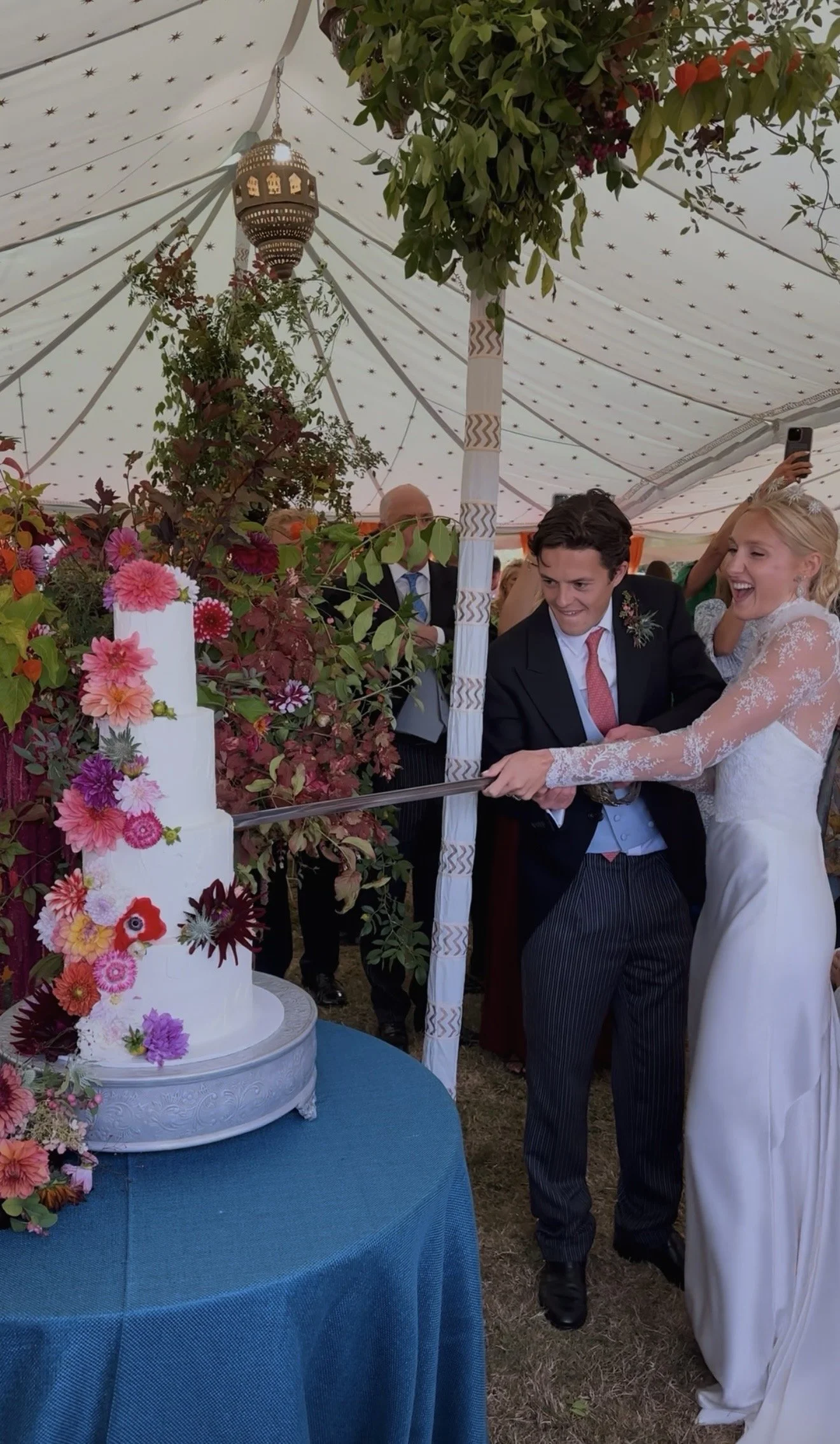 A bride and groom cutting a wedding cake at their wedding reception with a ceremonial sword under a decorative outdoor tent with hanging lanterns. The cake has 3 tiers and is heavily decorated with edible flowers.