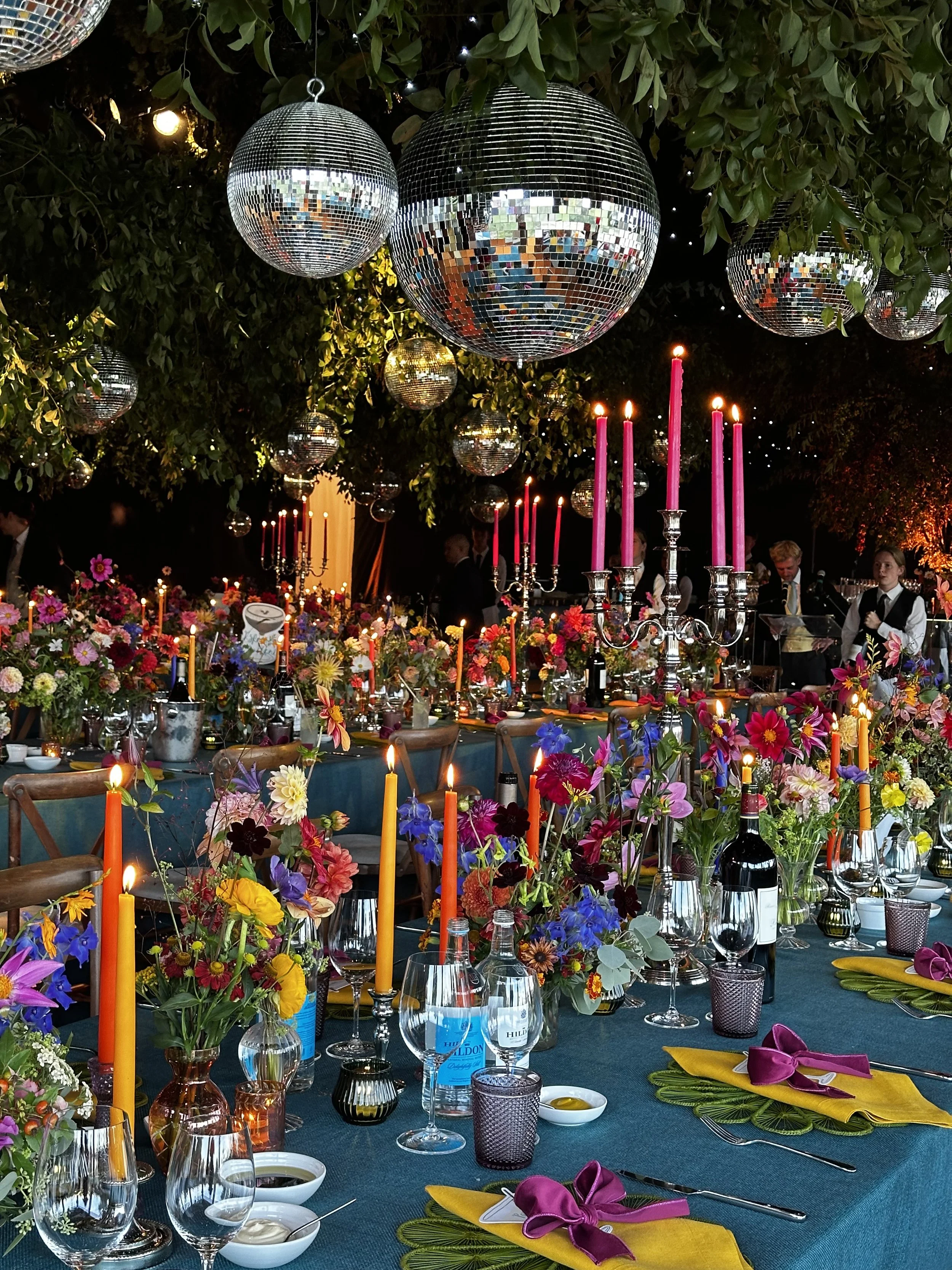 A heavily decorated indoor dining table at night with colorful flowers, tall bright candles, wine glasses, and large disco balls hanging from an indoor leafy canopy.