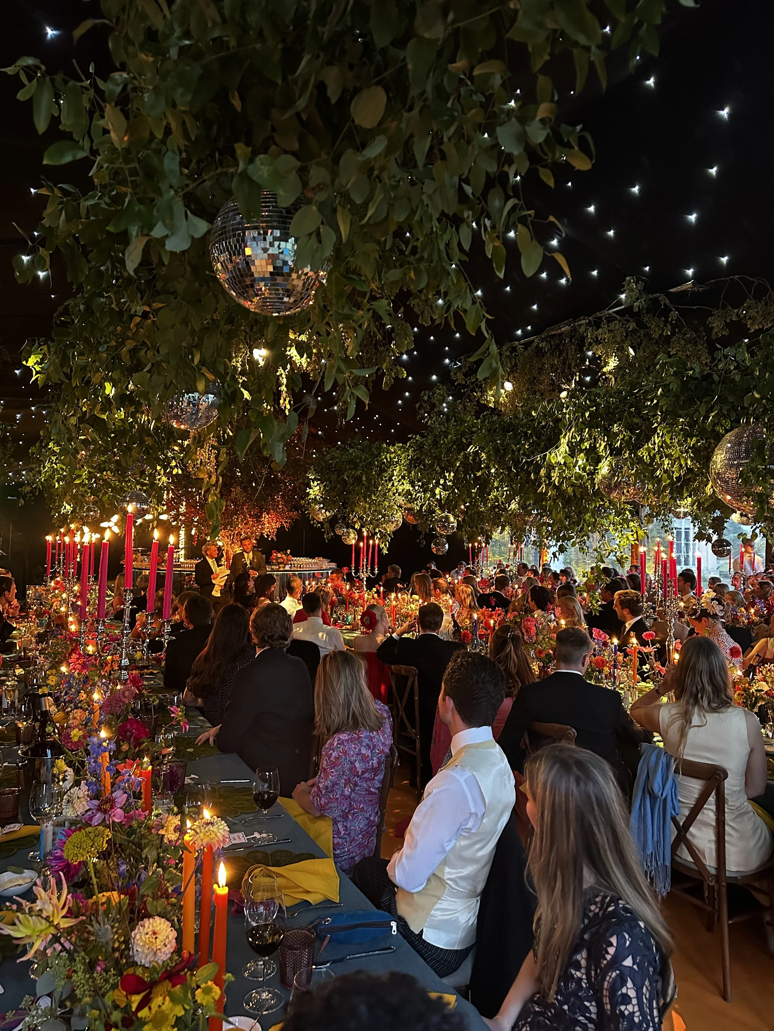 A large indoor banquet hall decorated for a celebration, with many guests seated at tables adorned with colorful flowers, candles, and tall pink candles. The ceiling has greenery and disco balls, and string lights are hanging overhead. There are two speakers and a person speaking at a small stage at the far end of the room.
