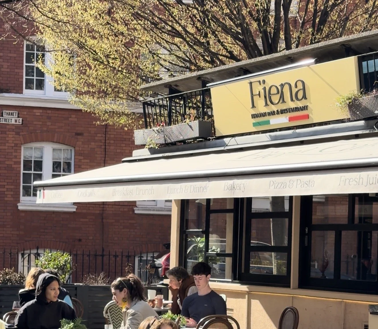Outdoor seating area of an Italian restaurant named Fiena, with customers dining under a awning with menu items printed on it, and a sign with the restaurant's name and Italian flag colors on top.