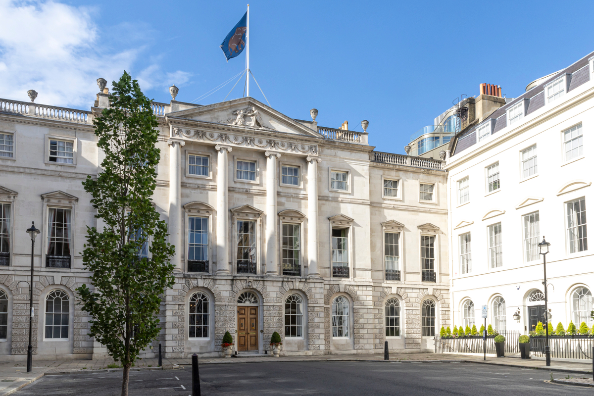 The historic Oriental Club building with classical architectural features, windows with decorative pediments, and a wooden front door, located on a city street under a blue sky with a navy flag displaying an elephant flying on top.