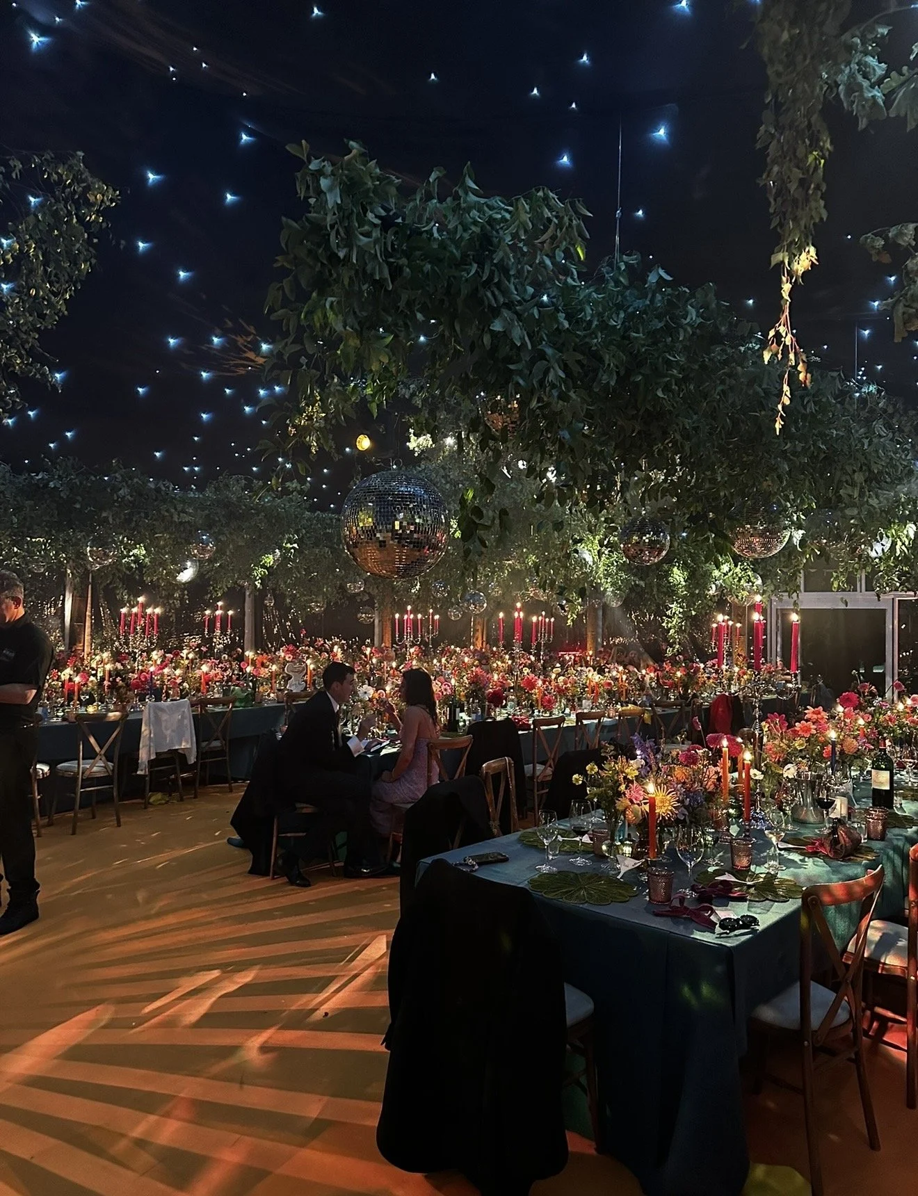 An elegant indoor event space decorated with numerous flowers, candles, and disco balls, with blue star-like ceiling lights, and people enjoying conversation.
