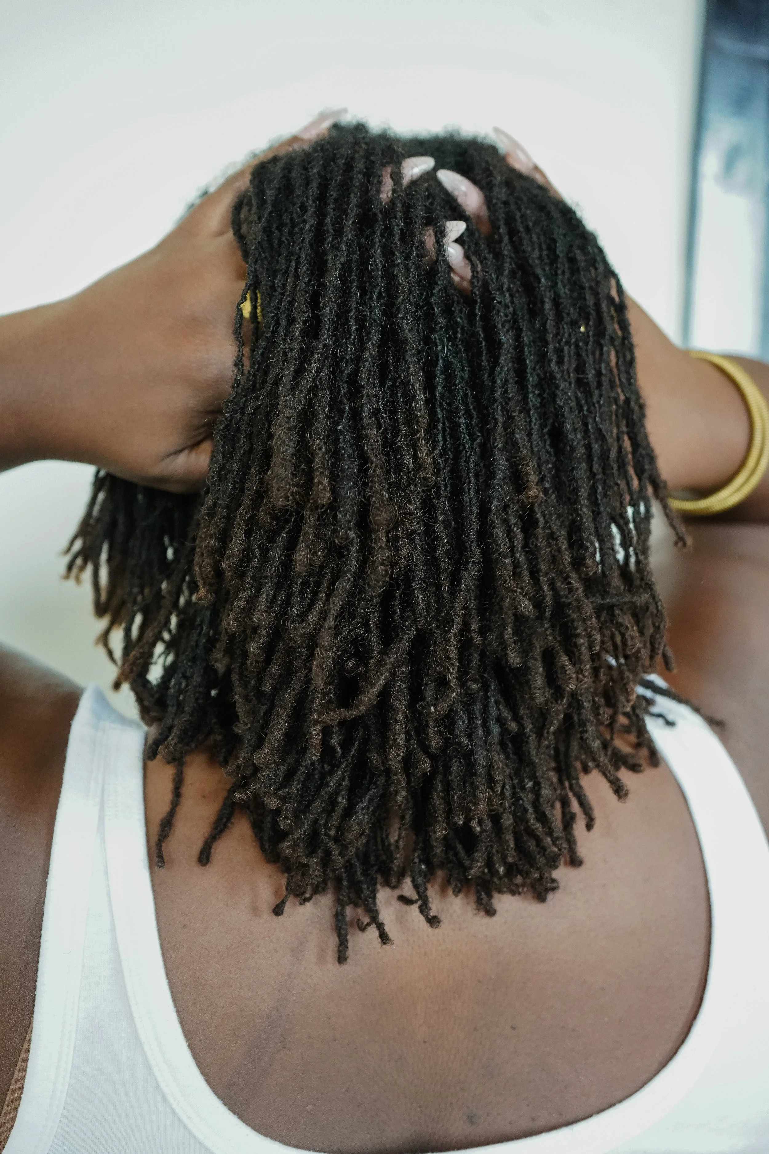 A person with shoulder-length fully matured micrlocs, wearing a white tank top and a gold bracelet, resting their hand on their head.