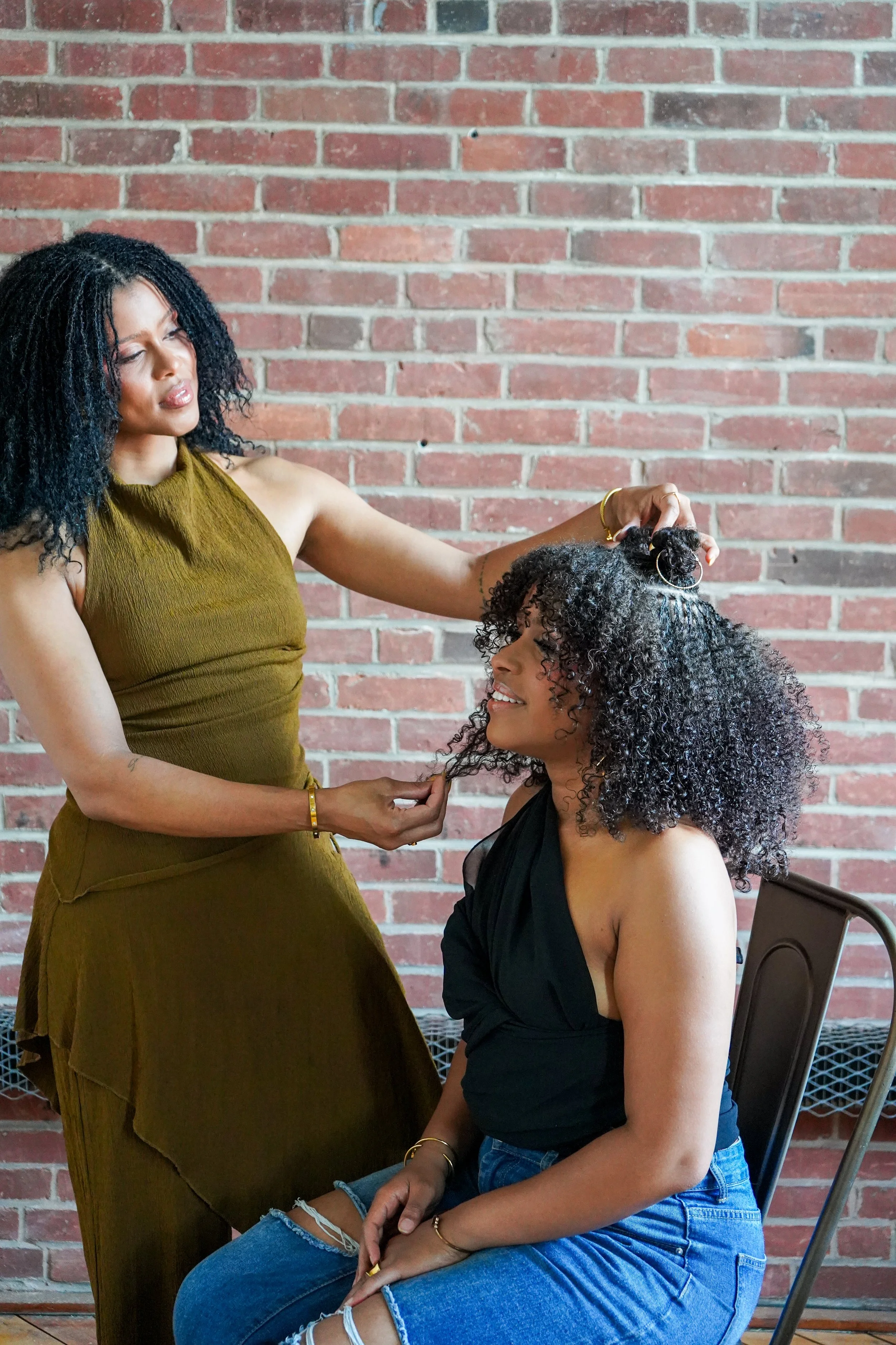 A client with curly hair and root braids is sitting in a chair, smiling, while Lorreta styles it. They are in front of a brick wall.