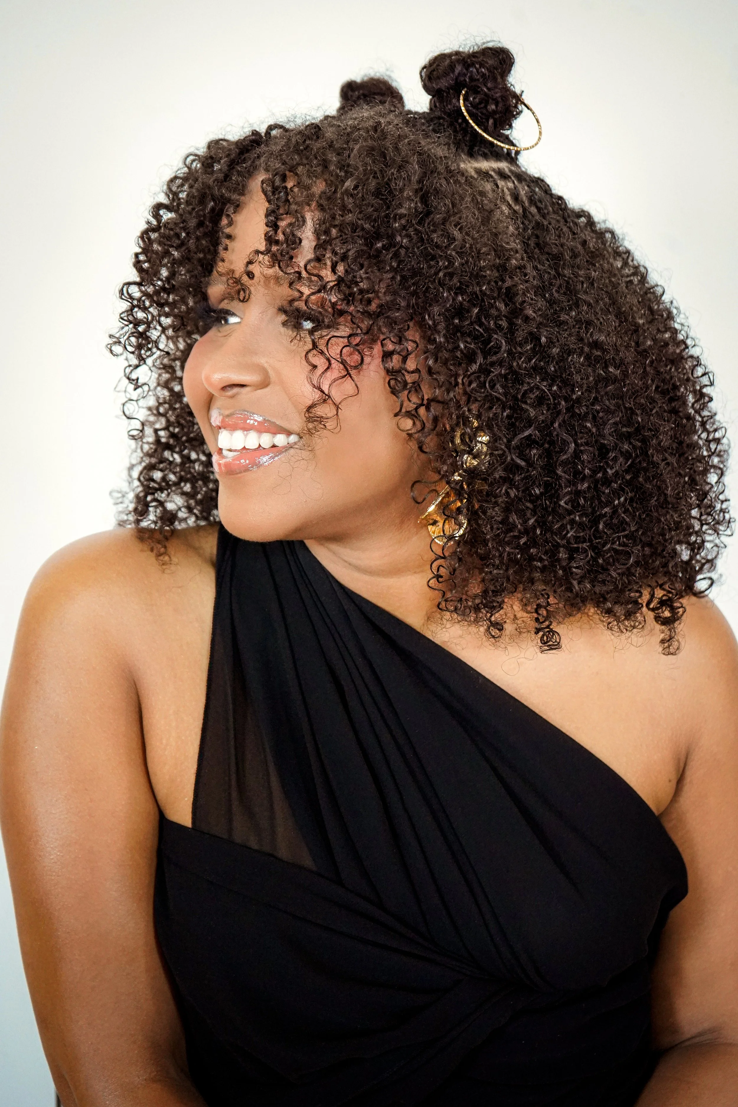 A woman with curly hair styled in two buns, wearing hoop earrings and a black, one-shoulder dress, smiling and looking to her left.