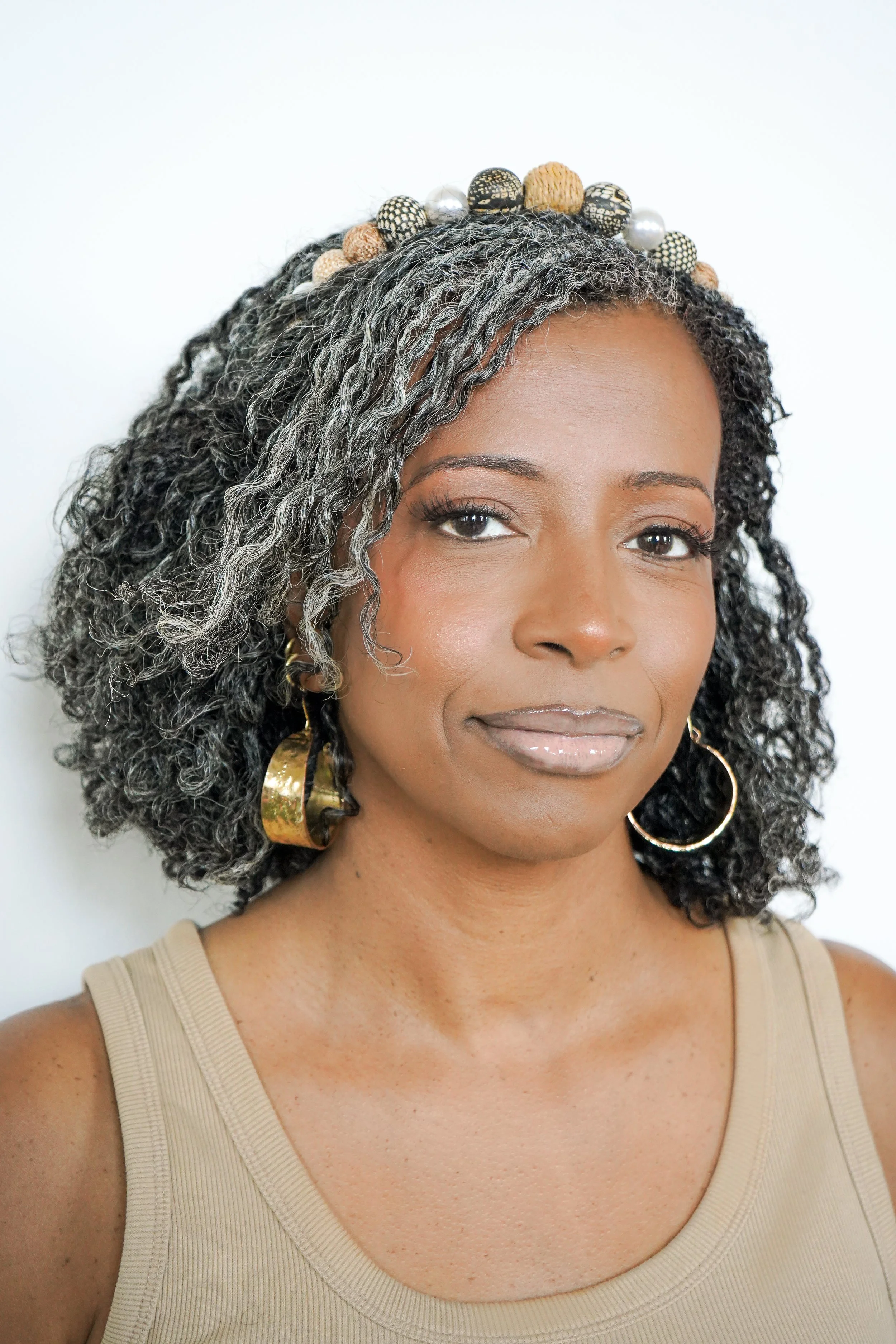 Close-up portrait of a woman with gray hair and newly installed micrlocs, wearing large gold hoop earrings, a beige sleeveless top, and decorative headband with black, white, and beige beads and pearls.